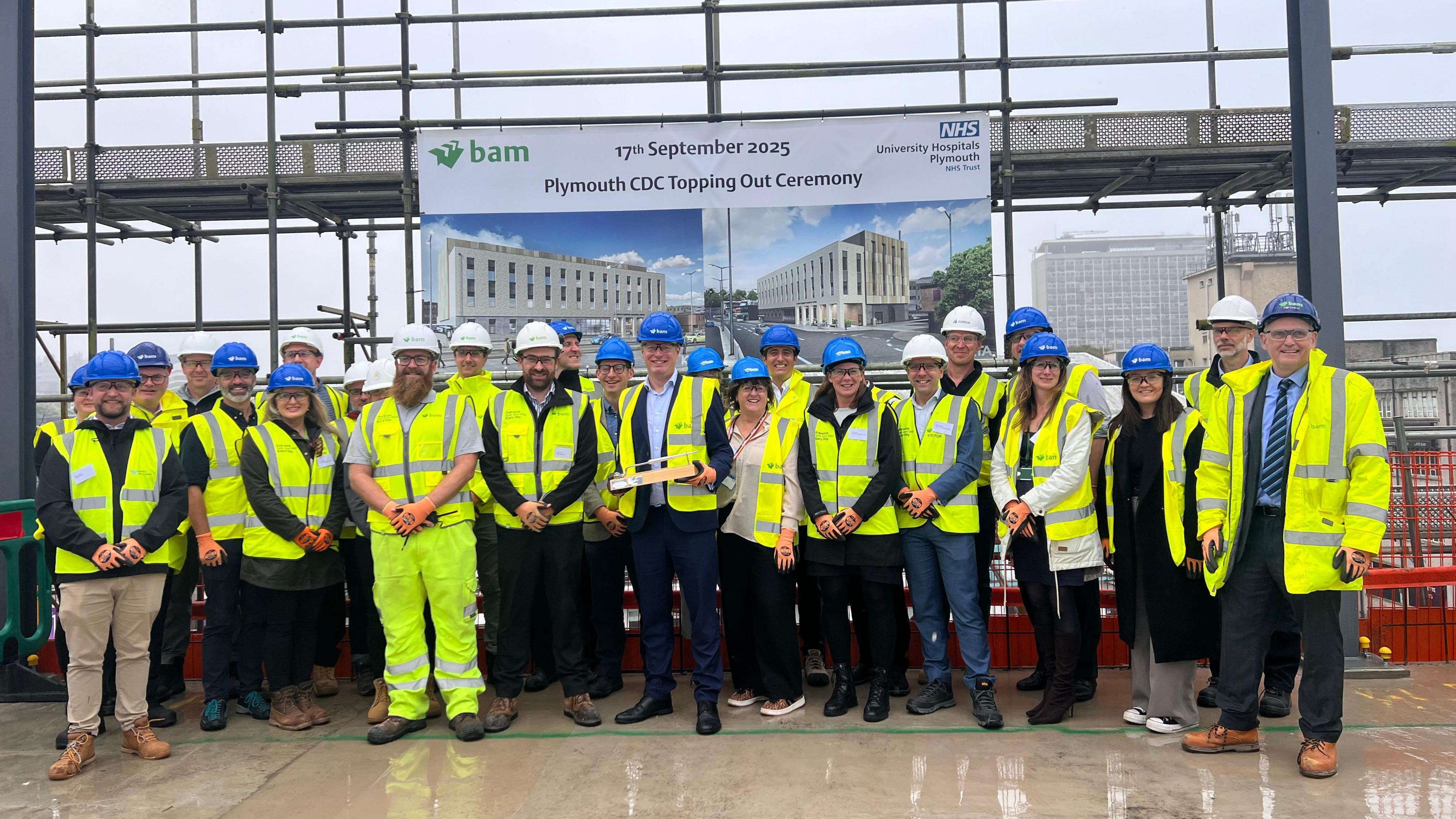 A group of workers wearing hi-vis at a construction building site. The group are standing in a row and are wearing blue hard hats. A banner displayed behind them says 17 September 2025 Plymouth CDC Topping Out Cermony.