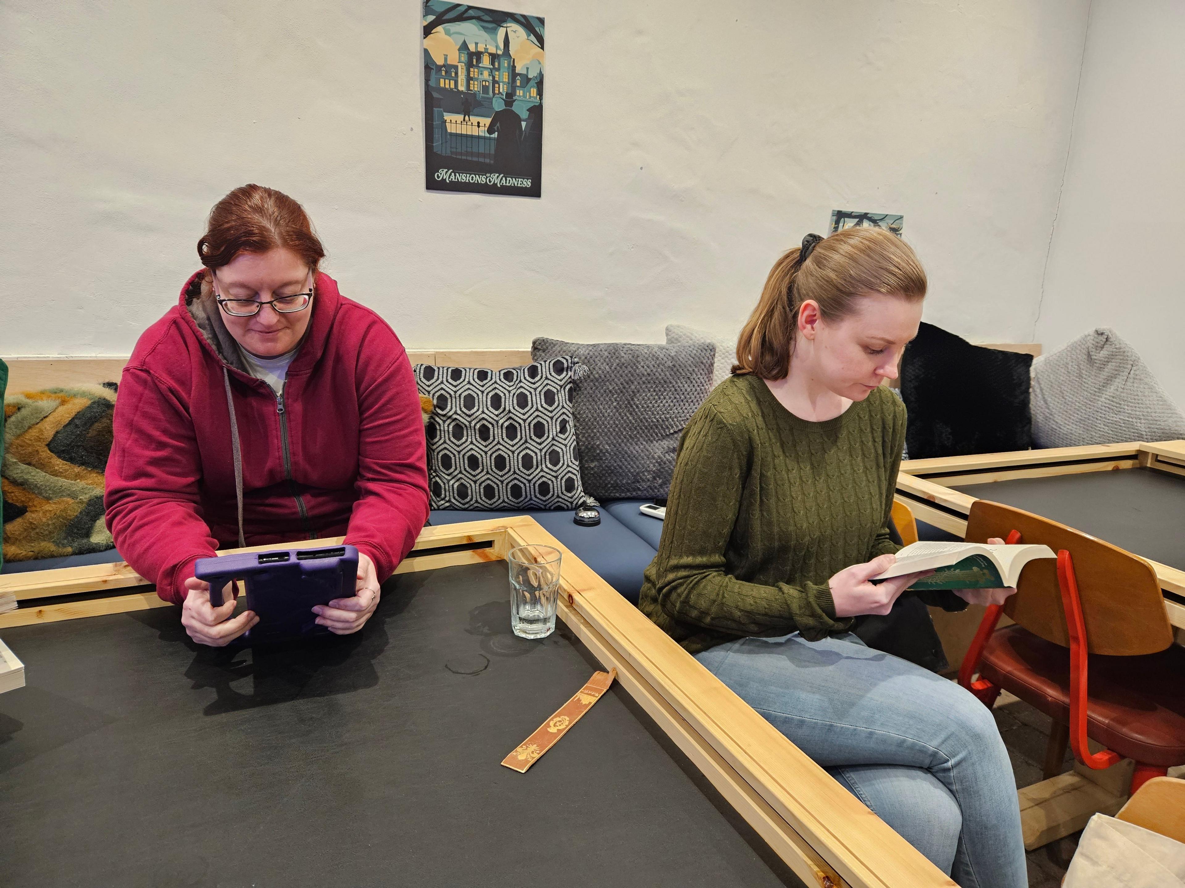Two readers of the silent book club read while sitting around a board game table. One, with reddish-brown hair, is reading from a purple tablet. The other, with lighter brown hair, is reading a paperback book.