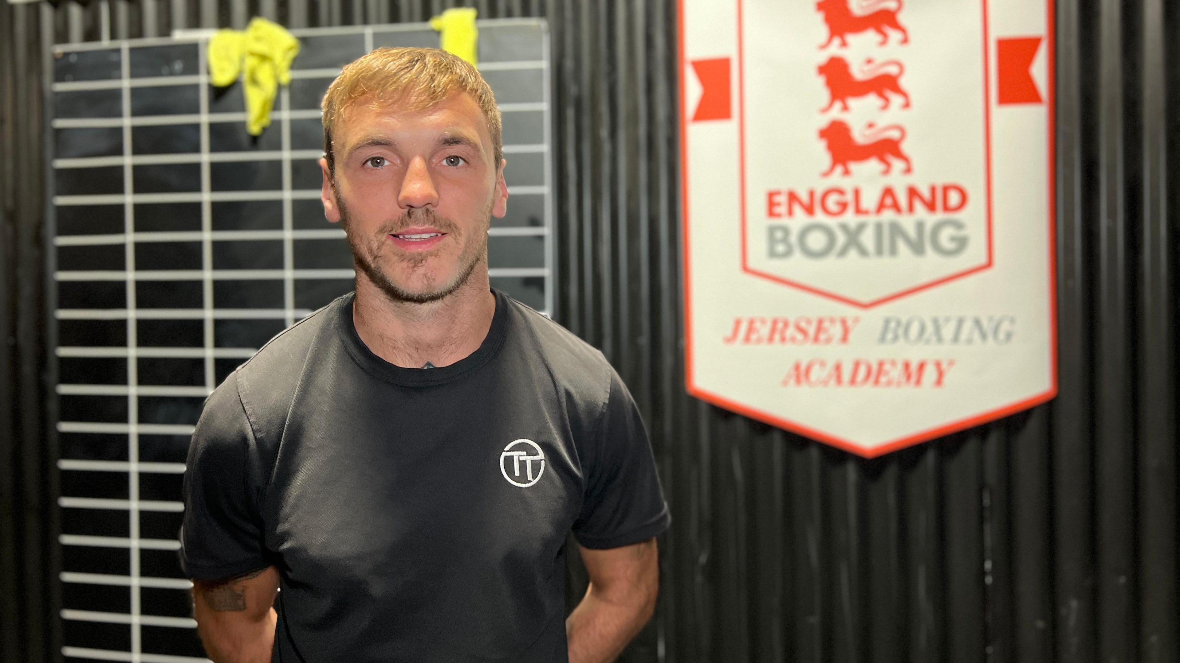 Tom smiles at the camera with his hands behind his back with an England Boxing sign behind him. He has short blonde hair and is wearing a black t-shirt