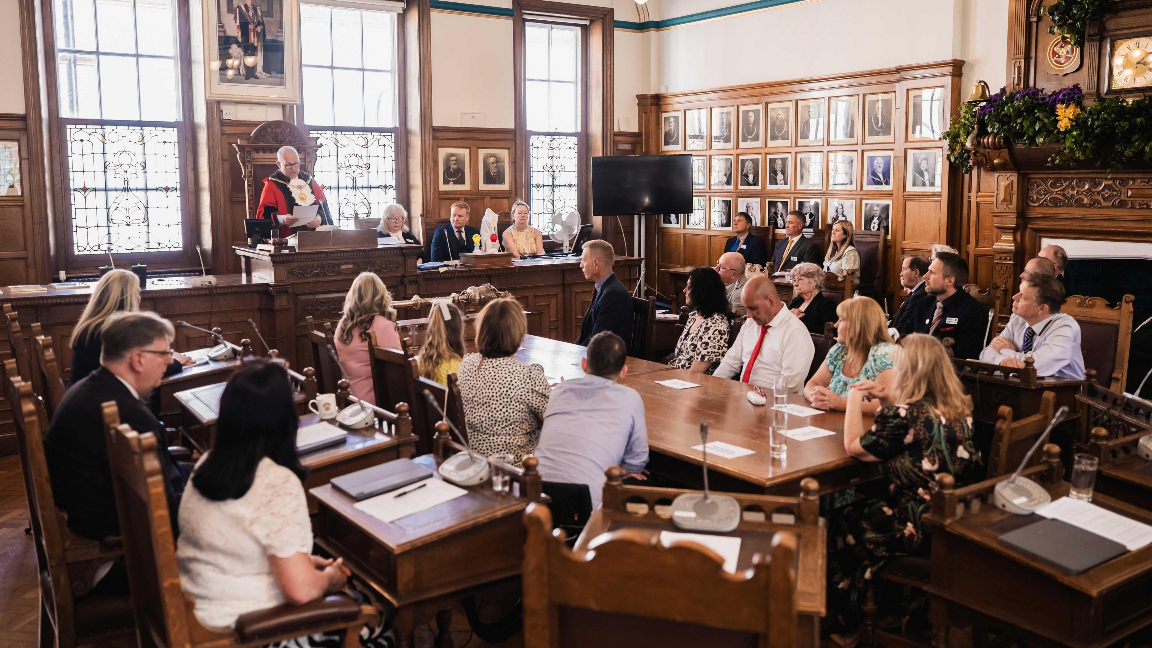 Steven Crellin makes his speech in the large wooden chamber, with people sitting around a large table and desks in the centre of the room.