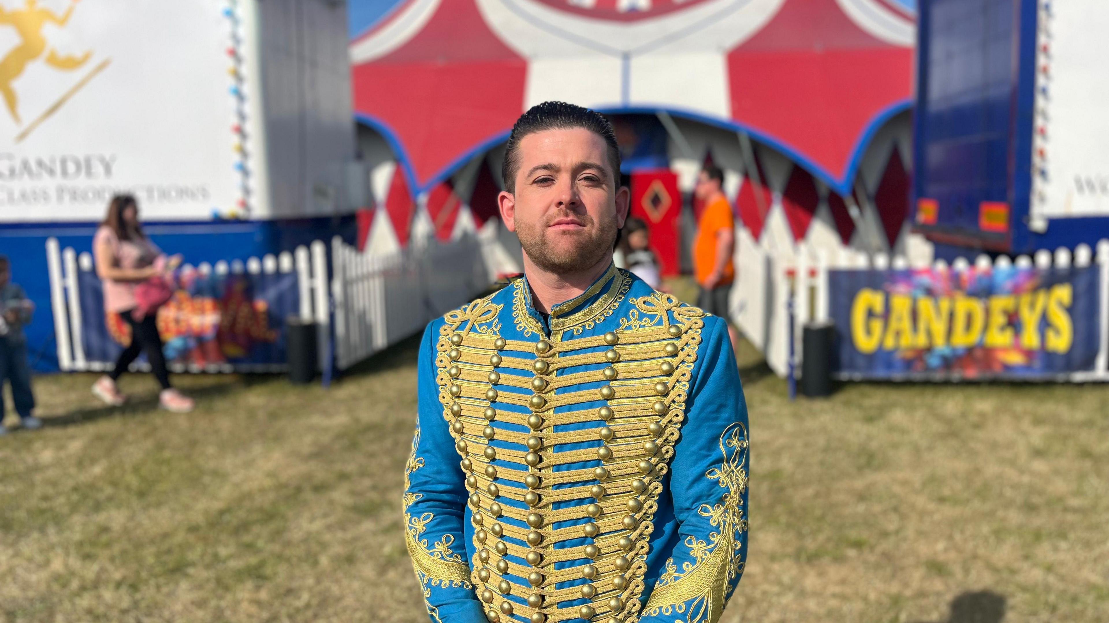 Joel Hatton, circus manager, faces the camera wearing a blue and gold circus jacket with buttons across the front. He looks serious but with a slightly wry smile.