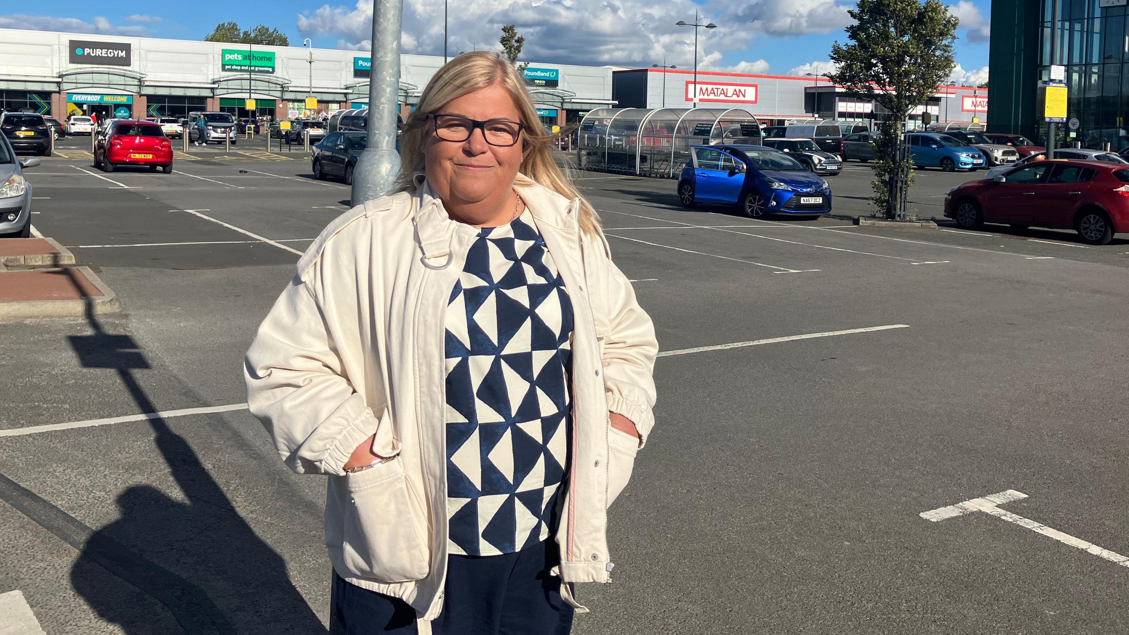 Councillor Kathryn Rooney at the car park. She wears a white jacket and a blouse with a navy blue and white geometrical print. She is blonde with glasses. Her hands are in the pockets of the jacket. 