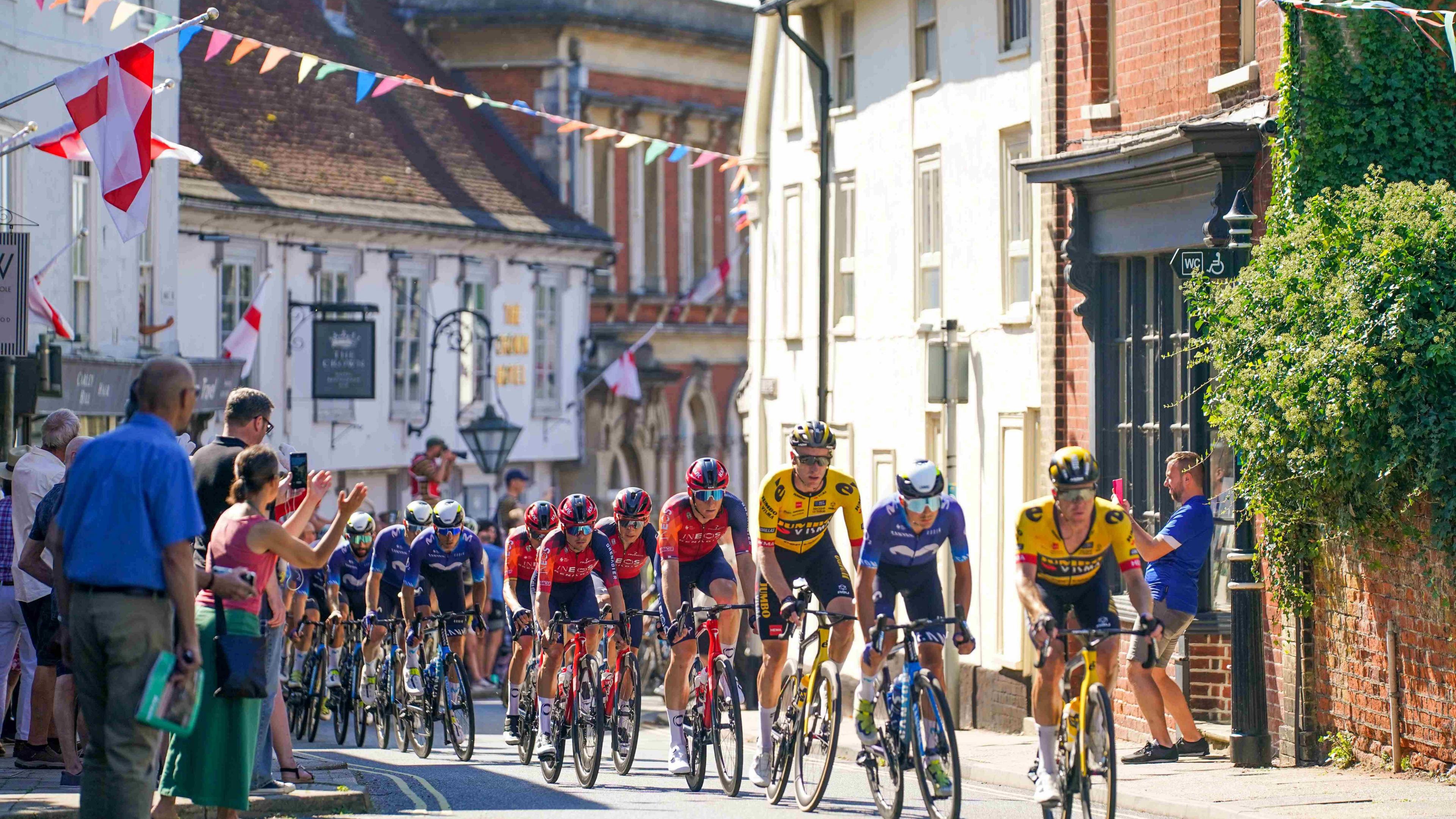 Cyclists ride through the streets of Framlingham. Spectators cheer them on from the pavements. Bunting is hung across the buildings either side of the road.