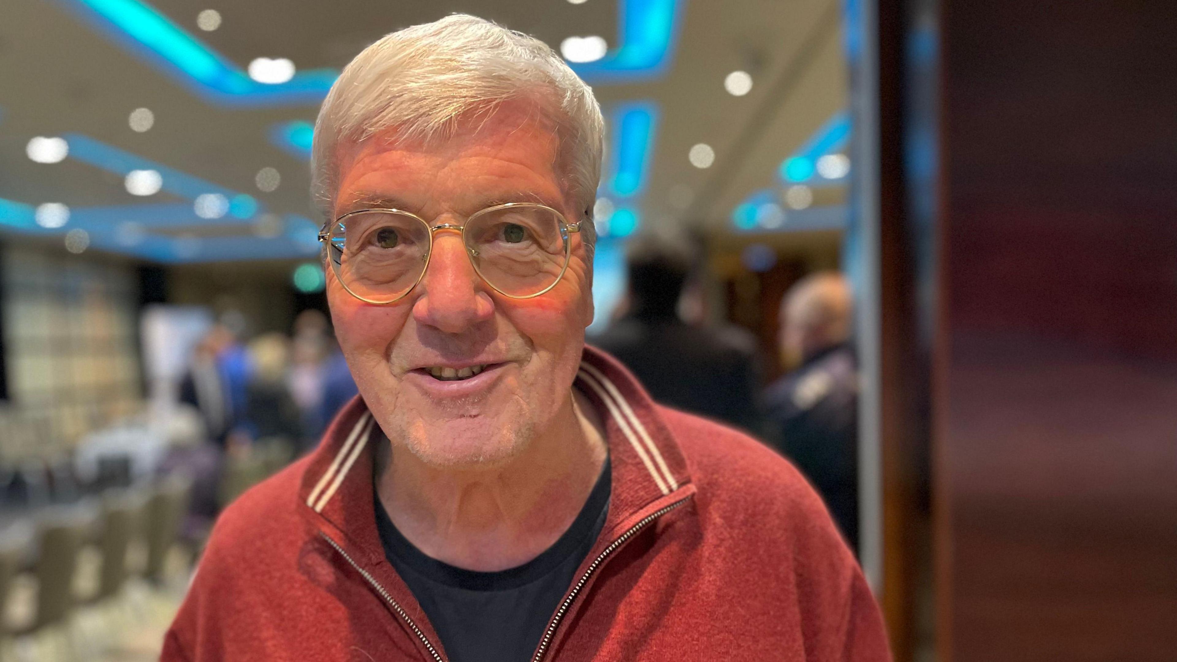 John Youngs is an older man with grey hair. He is smiling into the camera. He has a black t shirt and red jumper on, he is wearing large, round glasses with clear frames. The background is an out-of-focus meeting room.