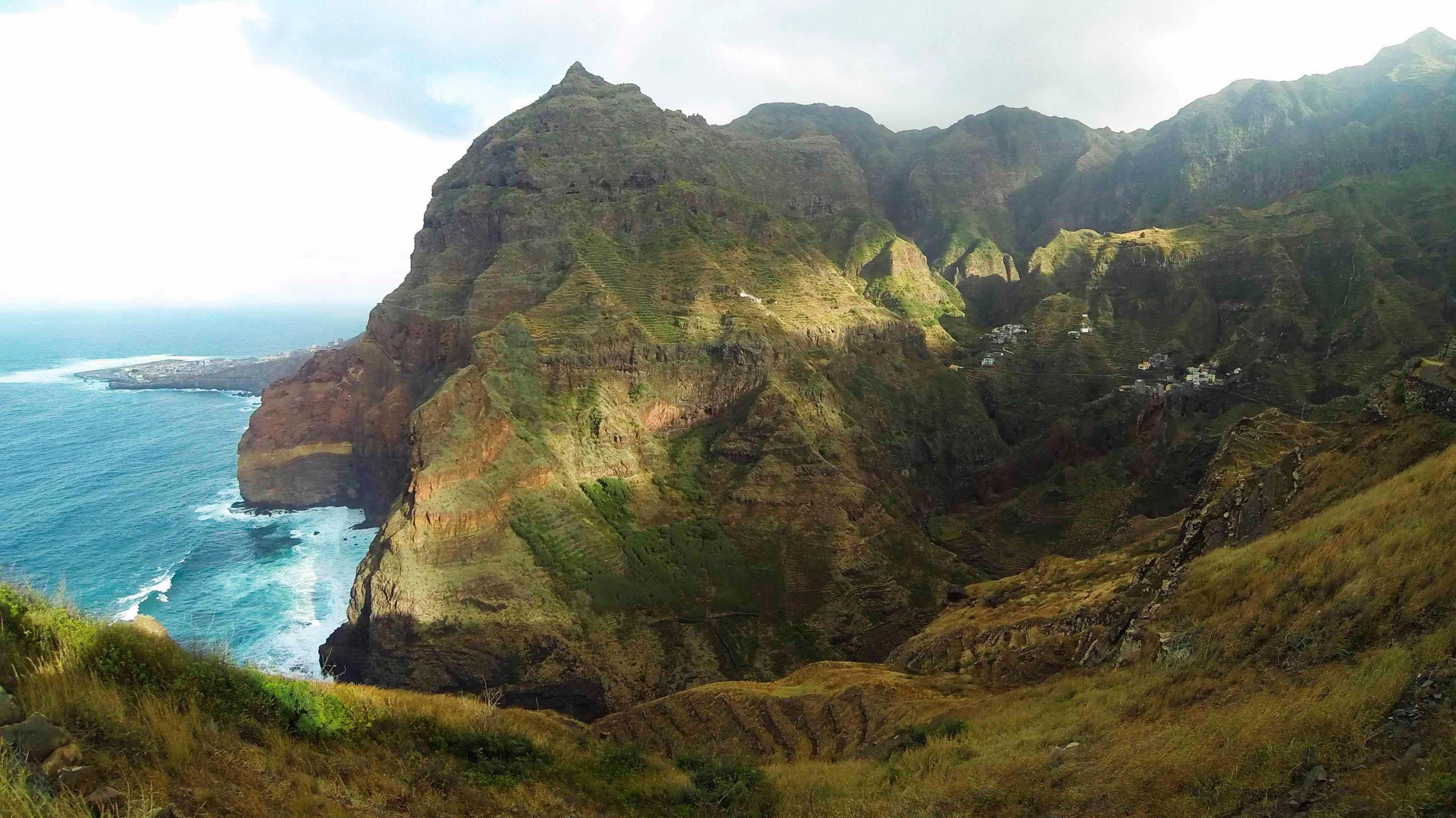 Foliage-covered green cliffs stand against the ocean on the northern coast of the island of Santo Antao, Cape Verde. Below is a bright blue sea. There are waves crashing against the cliffs.