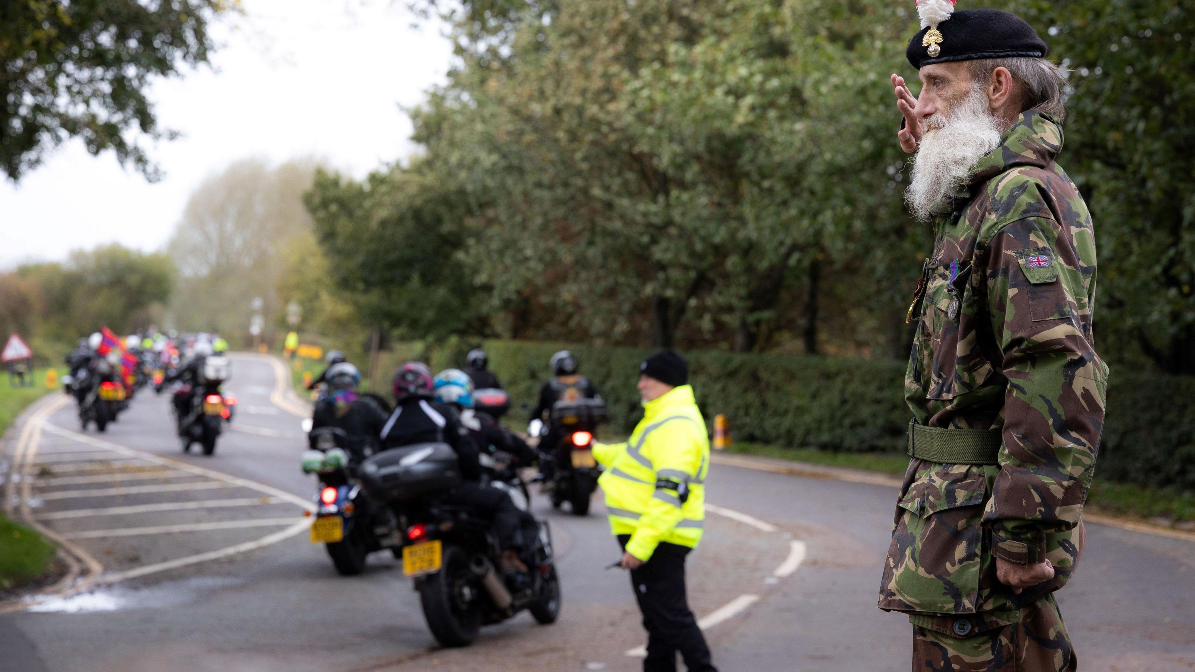 A large group of motorbikes on a road. A steward in a green high visibility jacket is on the road. At the front of the image a man wearing a uniform with a camouflage pattern who is saluting the motorcyclists. He is wearing a beret and has a grey beard and dark grey hair.