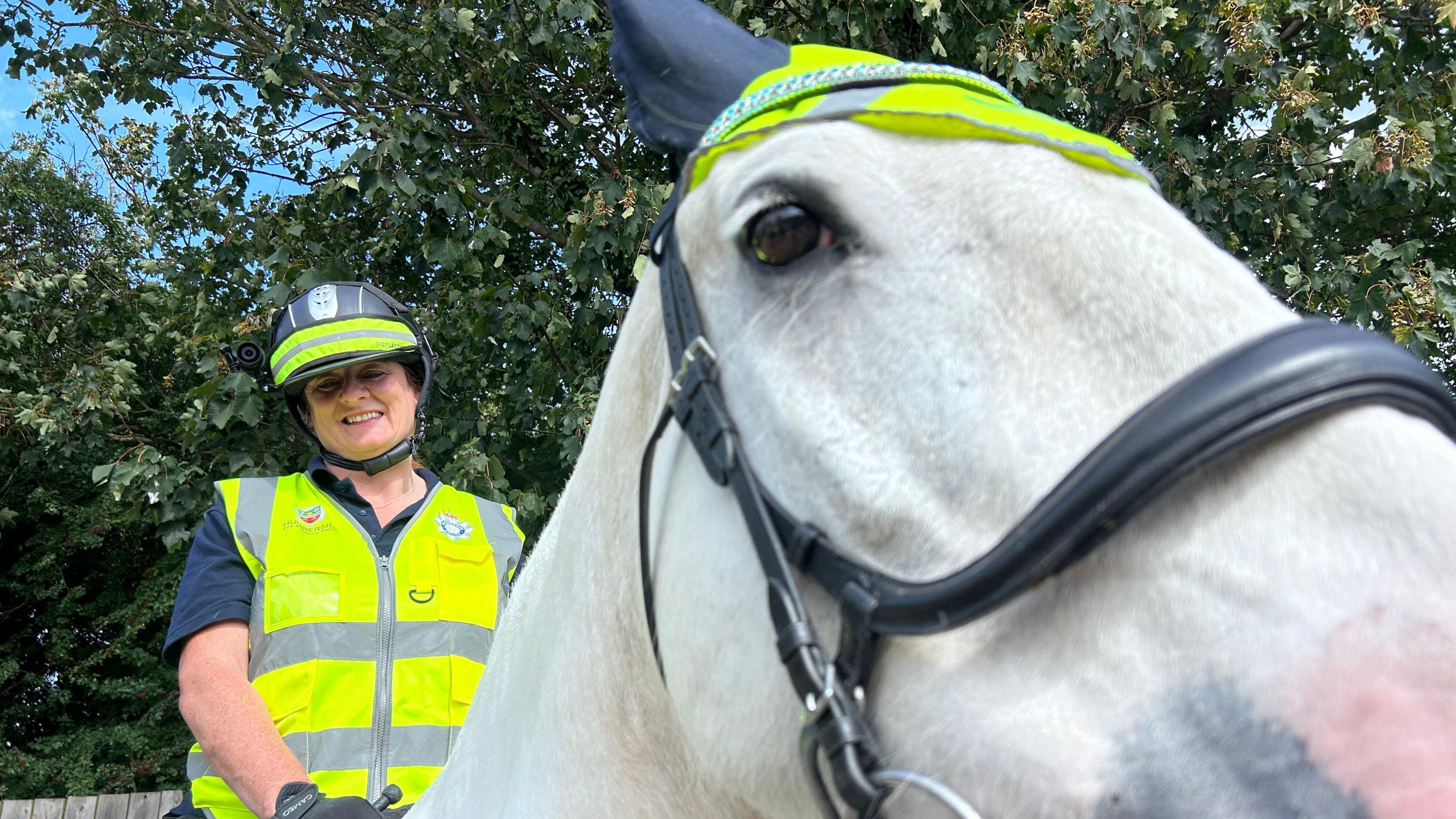 A woman wearing a high vis waist coat with a black helmet on that has a high vis strip around it, she's wearing a head cam pointing towards the camera and is sat on a white horse holding the reins. The horse has a sparkly head band around its ears and a small high vis hat on with his left eye looking towards the camera.