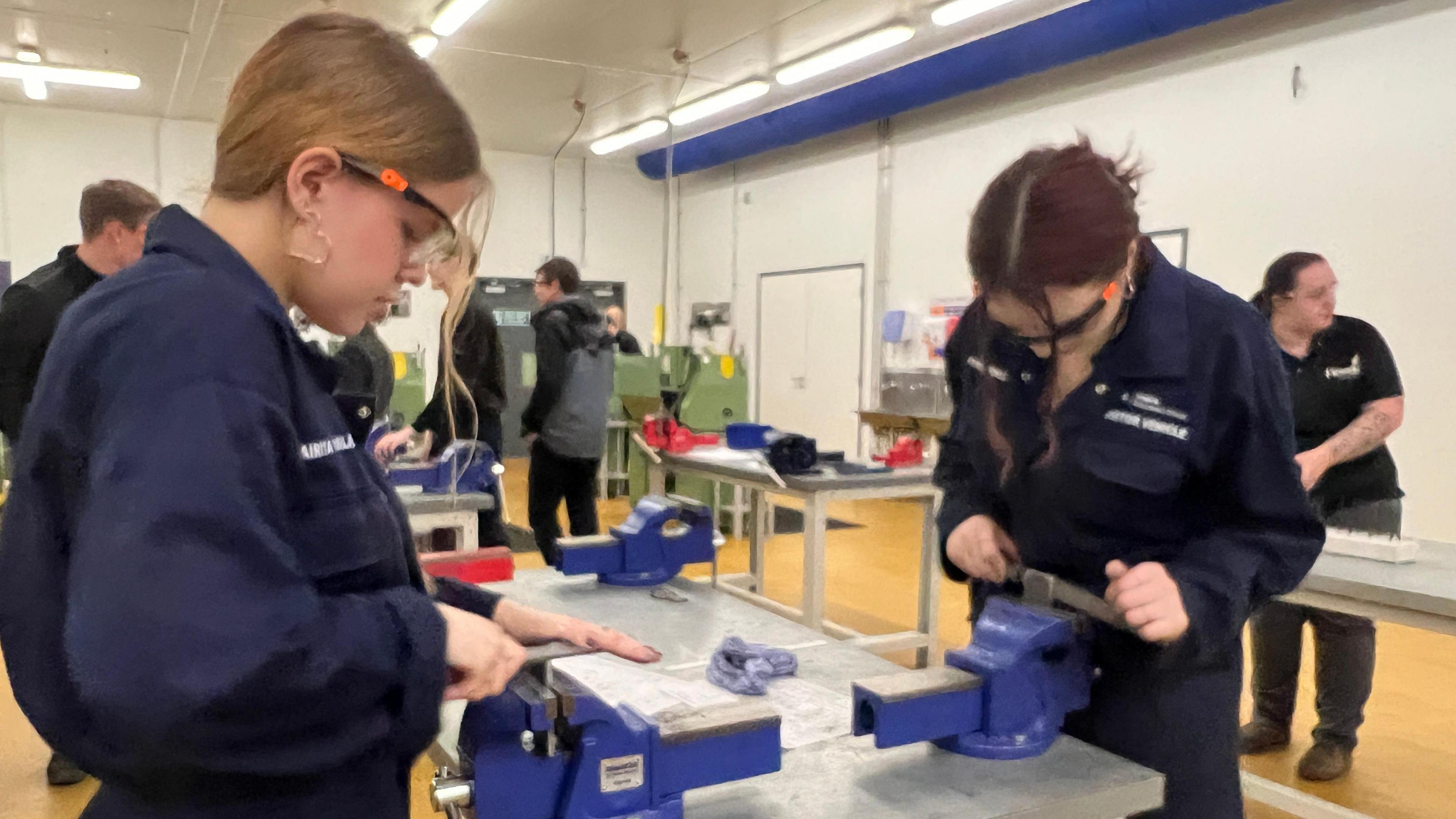 In a workshop two female apprenticeships wearing dark blue overalls are working with bench vices, in the room there are several benches