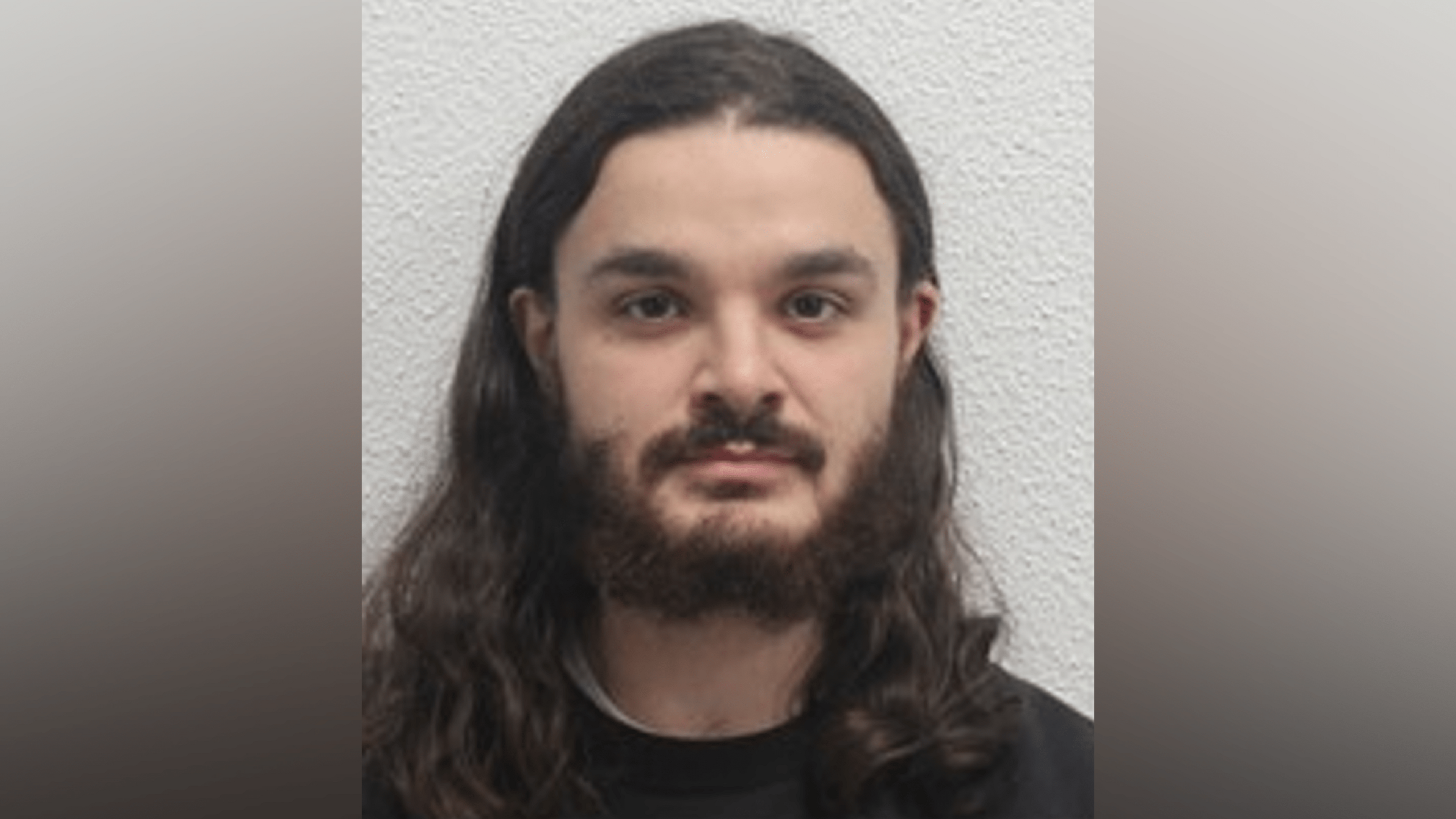 Declan George Candiani custody photo. He has long dark hair and a beard and moustache. He wears a black top. 