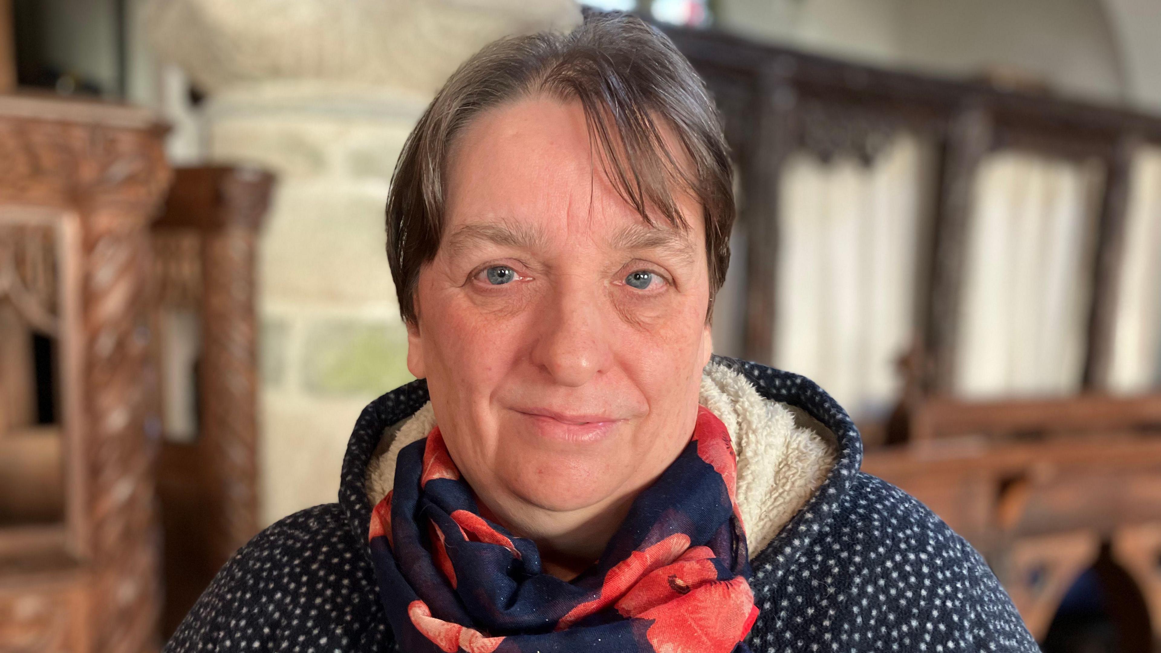 Sara Burrows pictured at the church. She is wearing a blue jumper with white dots and a fleece like hood. She is wearing a scarf with a poppy design on it.