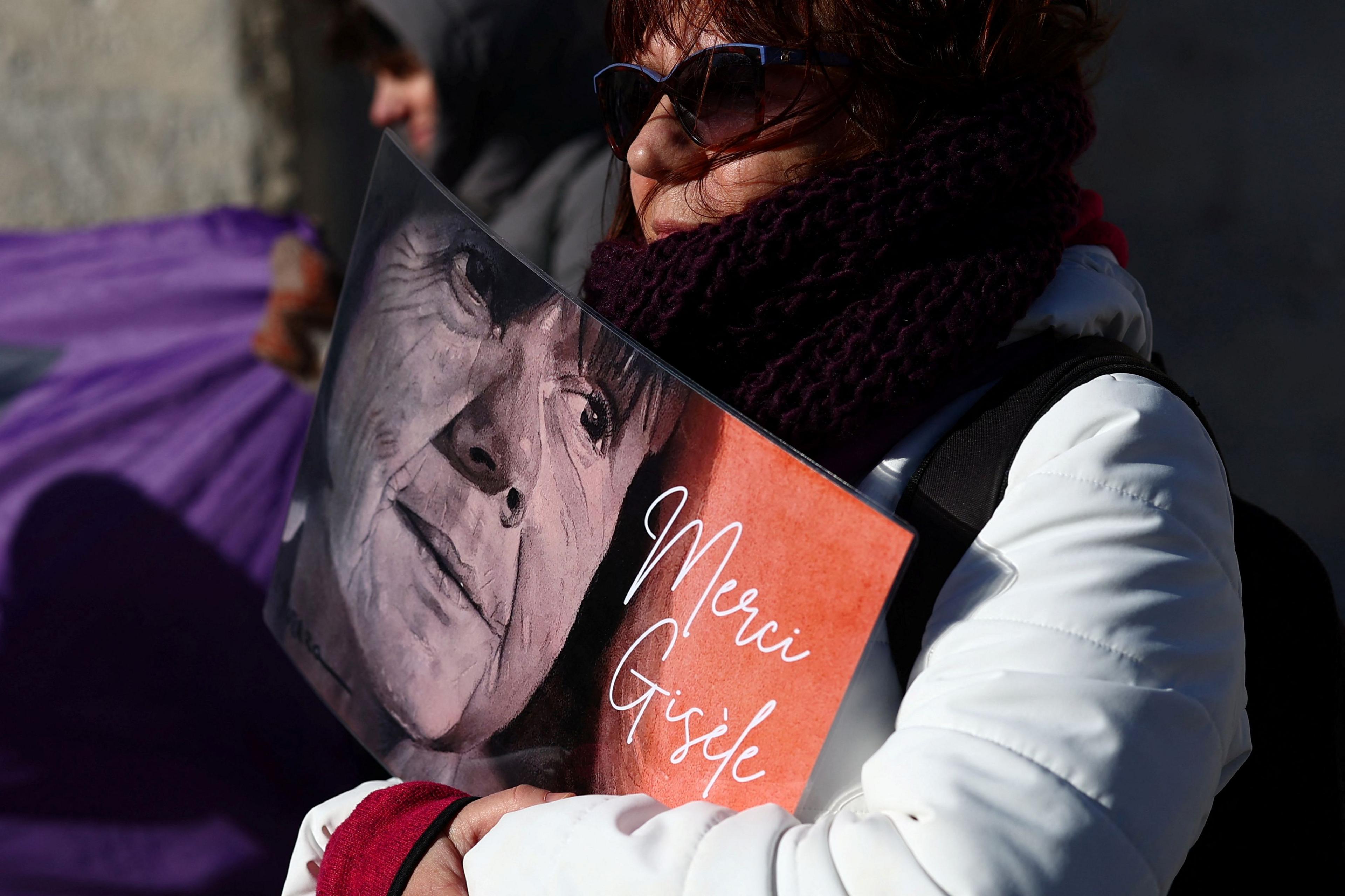 A woman wearing a white coat and sunglasses holds a sign with Gisèle Pelicot's face with the words "merci Gisèle".