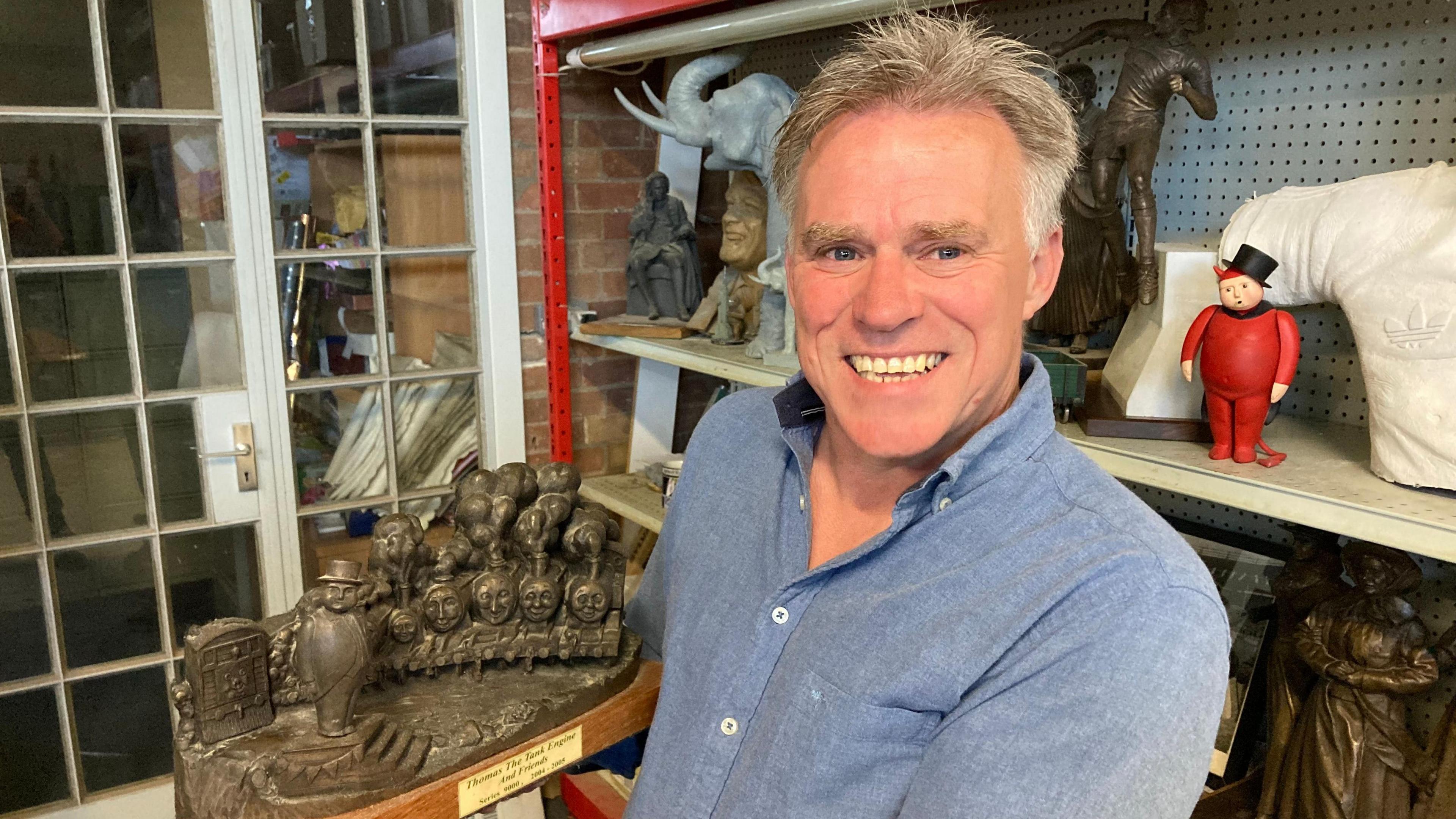 Sean Hedges-Quinn holds a sculpture as he smiles at the camera. The bronze sculpture shows several of the Thomas & Friends characters along with the Fat Controller. Mr Hedges-Quinn has short grey hair and wears a blue shirt.