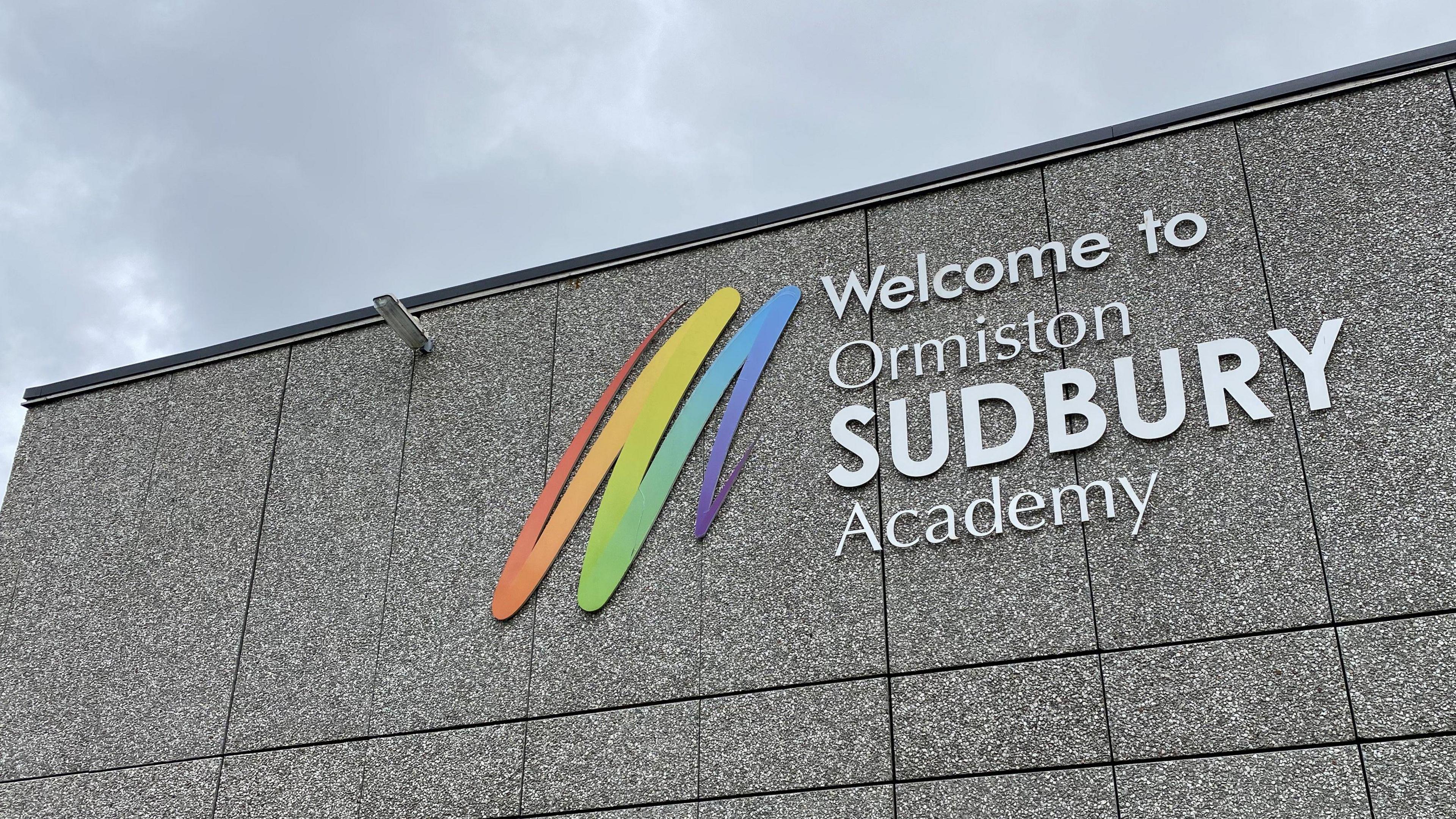 A general view of the exterior of Ormiston Sudbury Academy. The grey coloured building has the name of the school written on it white writing that reads 'Welcome to Ormiston Sudbury Academy'.