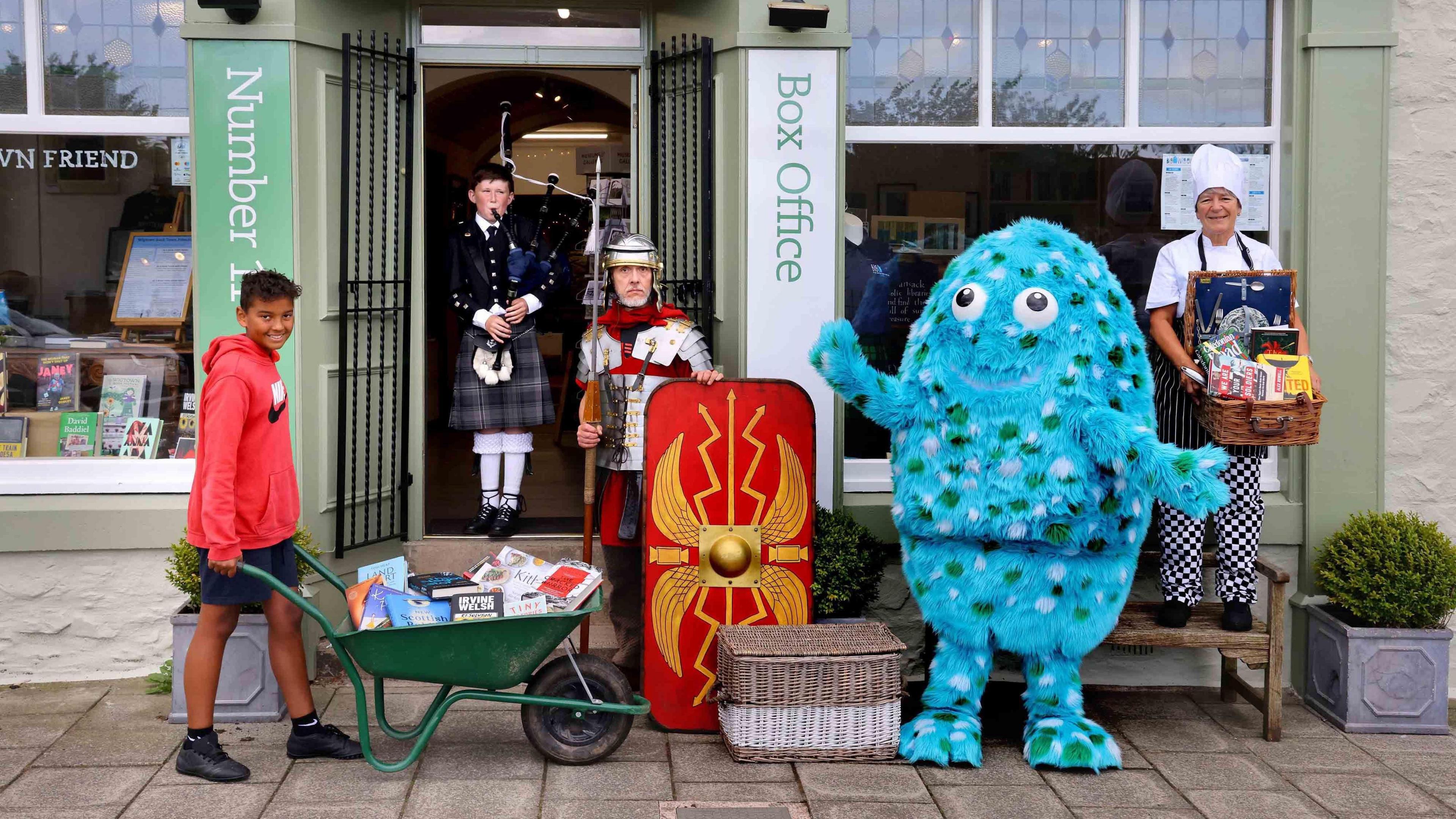 A boy with a wheelbarrow full of books, someone dressed up as a blue monster and a Roman soldier pose for a photograph outside a doorway with a box office sign beside it