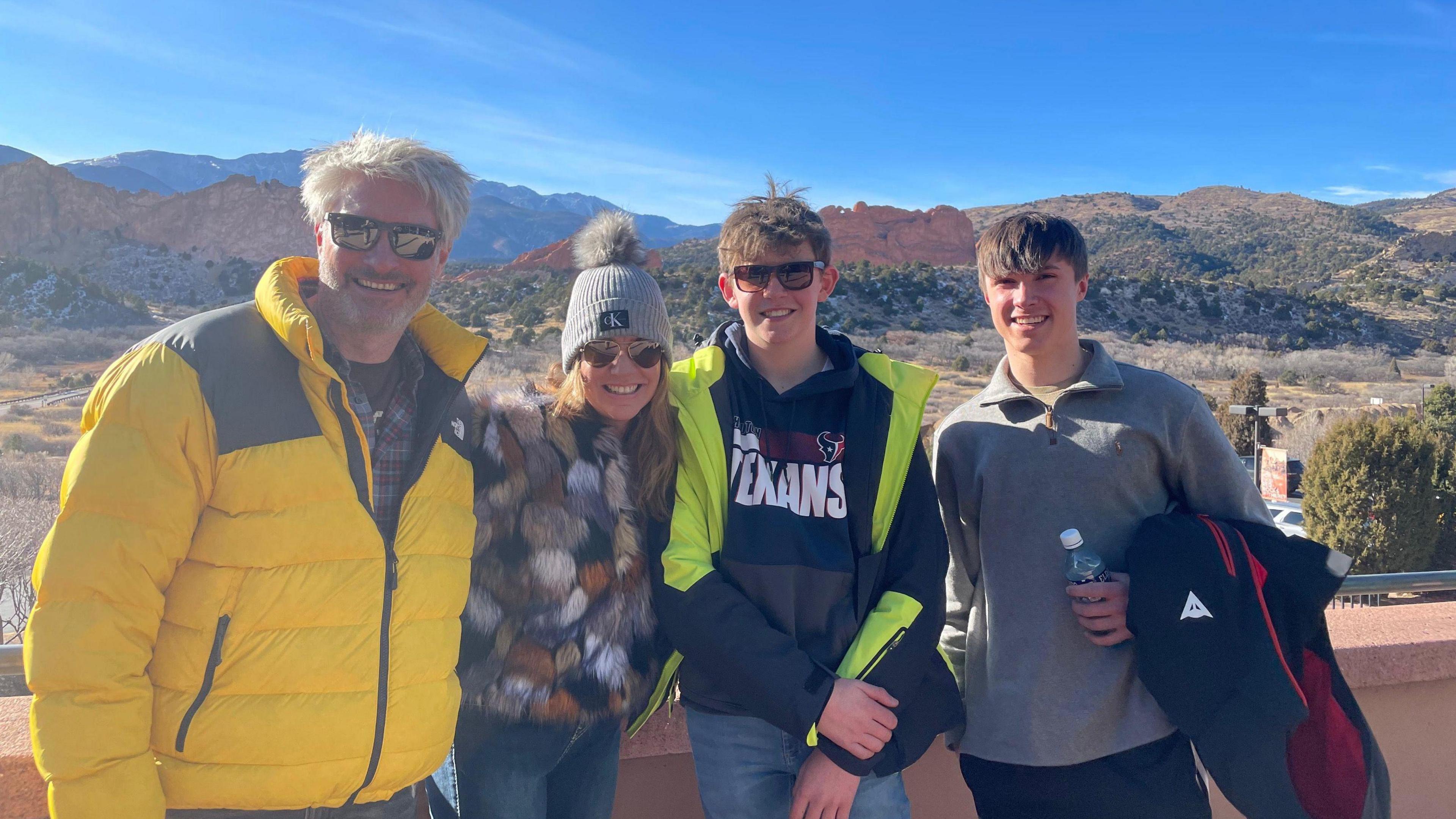 Family photo of the Webbers in front of a mountain view with a blue sky behind. The family is wearing warm coats.