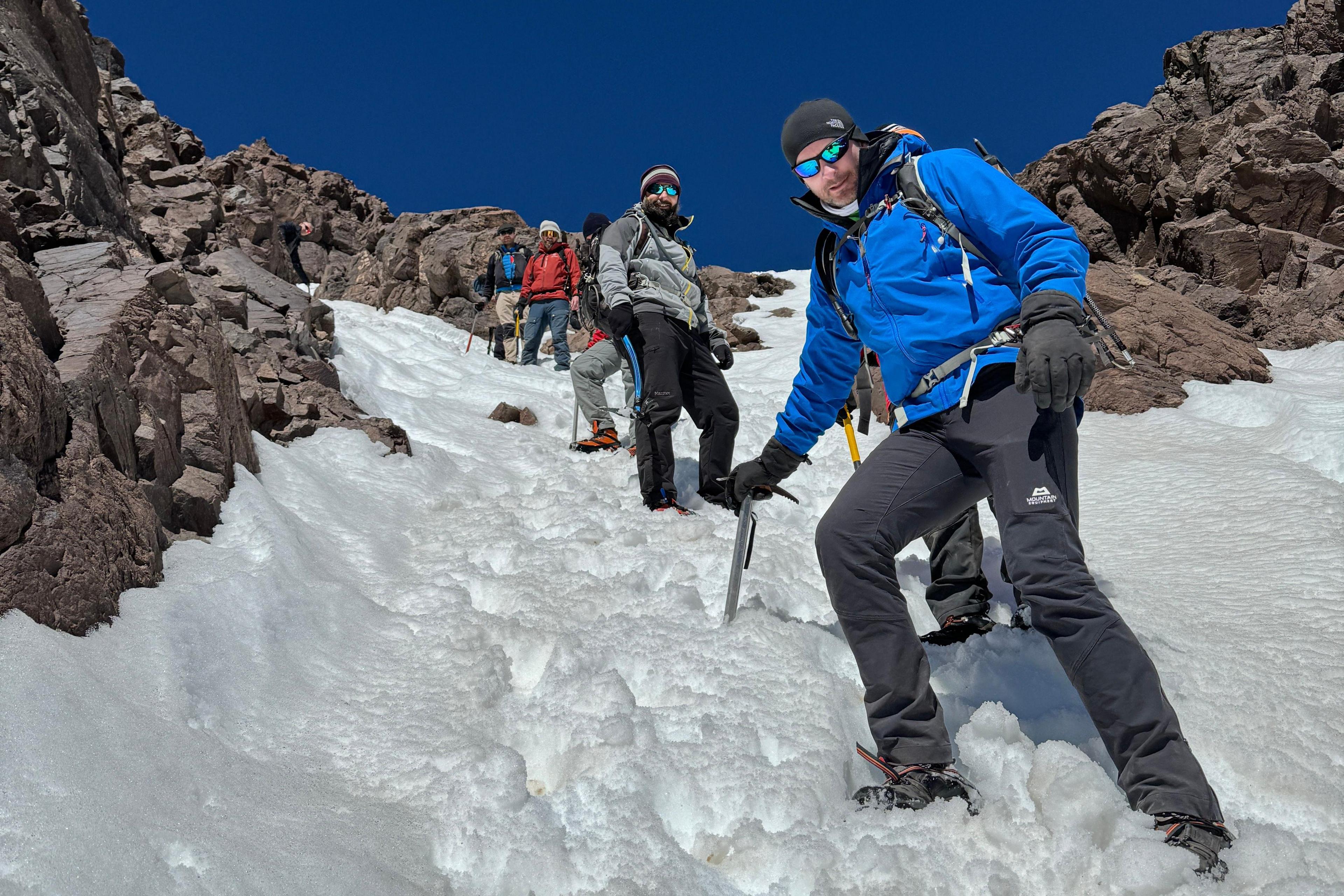 The photo is taken on the side of a snowy mountain, with some jagged rocks on show, and a deep blue cloudless sky. The photo has been taken from below with Gerwyn stood in the foreground of the photo, and five other men visible to varying degrees in the background. Gerwyn is leaning on an ice axe which has been wedged in the snow. Every man is wearing warm winter gear and sunglasses. 
