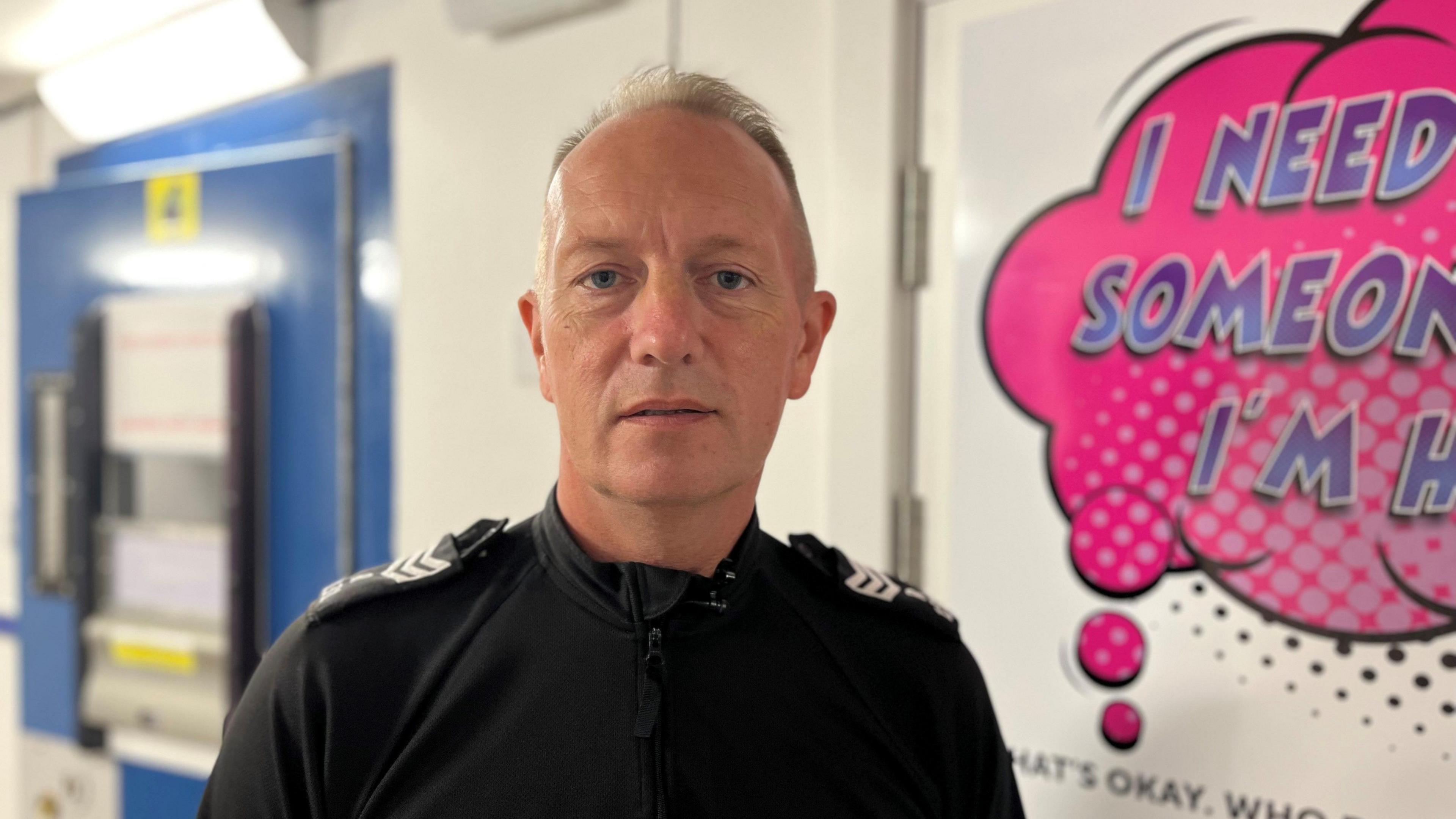 A photo of Custody Sgt Derek Taggart. He is in the police station looking directly at the camera. He is stood by a wall which carries a speech bubble in pink but the message is partially obscured. He is wearing a black police shirt.