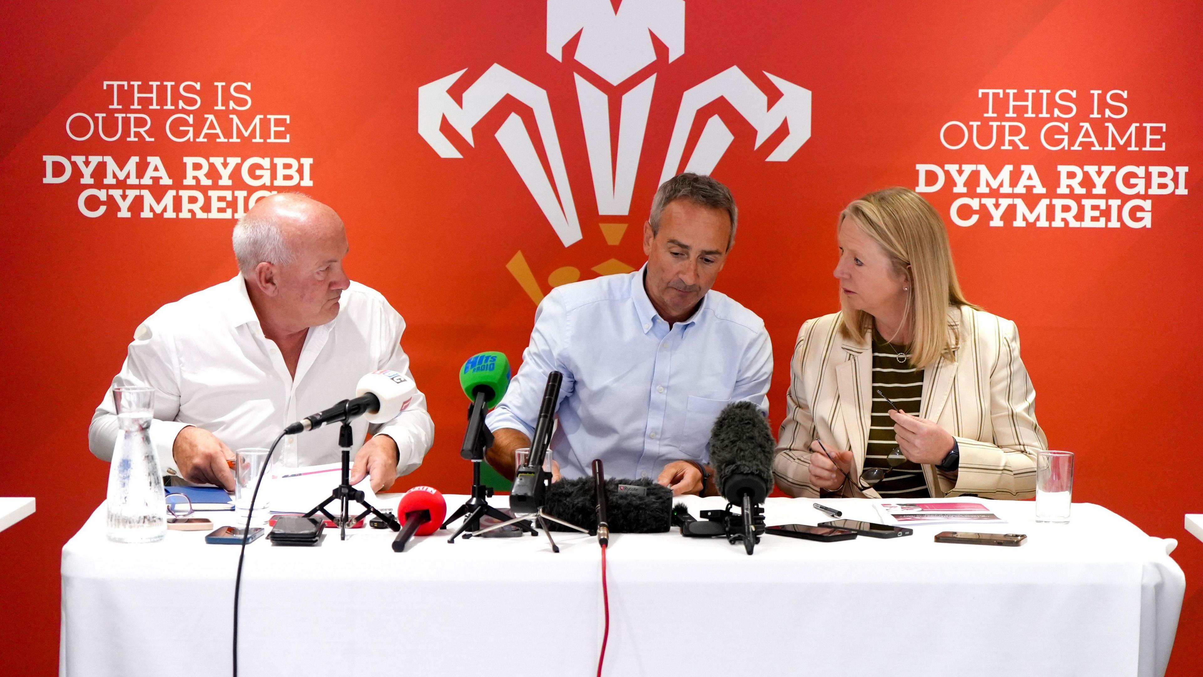 Richard Collier-Keywood, WRU chairman, Dave Reddin, WRU director of rugby and elite performance and Abi Tierney, WRU chief executive speaking at a press conference at the Principality Stadium in Cardiff. They are sat behind a white table with a WRU red background behind them. On the table is microphones, phones and a jug of water.