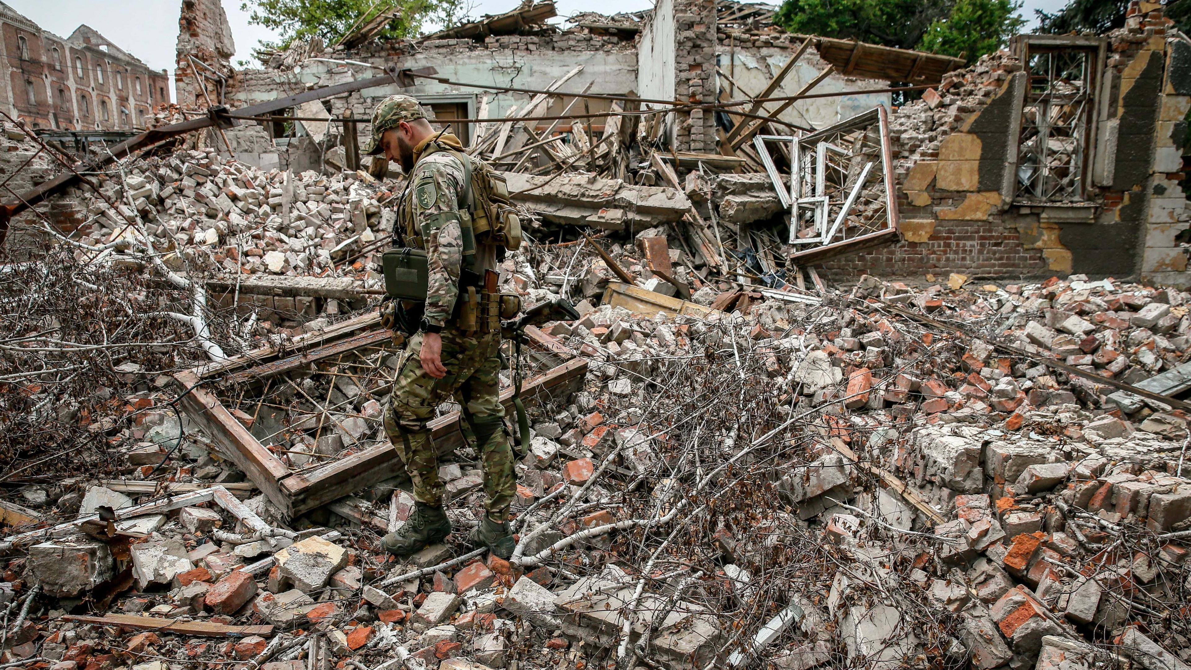 A Ukrainian soldier stands on large piles of rubble where a house once stood. There are bombed buildings in the background