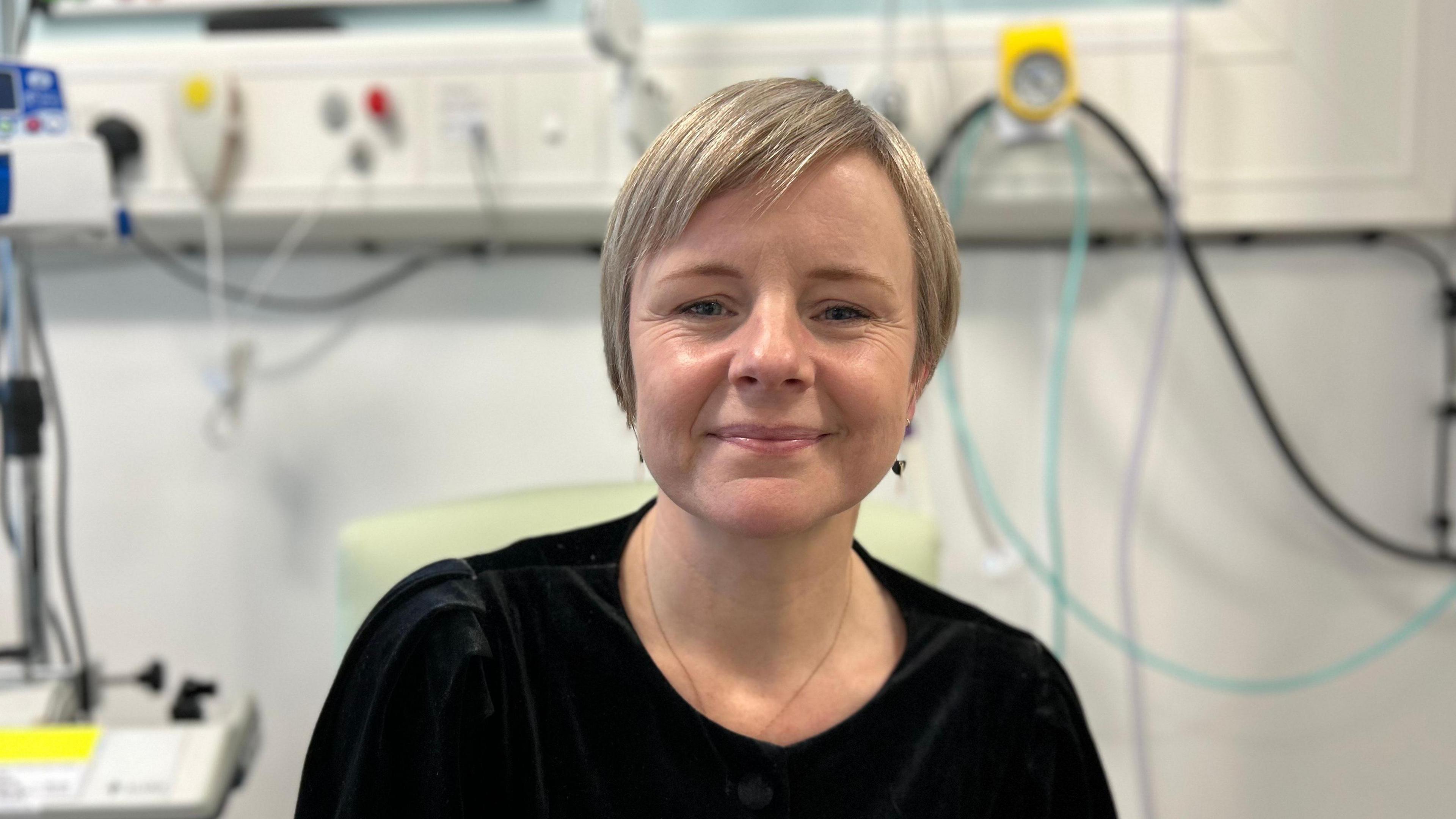 A woman with short blonde hair, ear rings and a necklace and a black top is sitting in a hospital environment. There are an assortment of different coloured tubes and a technical machine out of focus behind her.