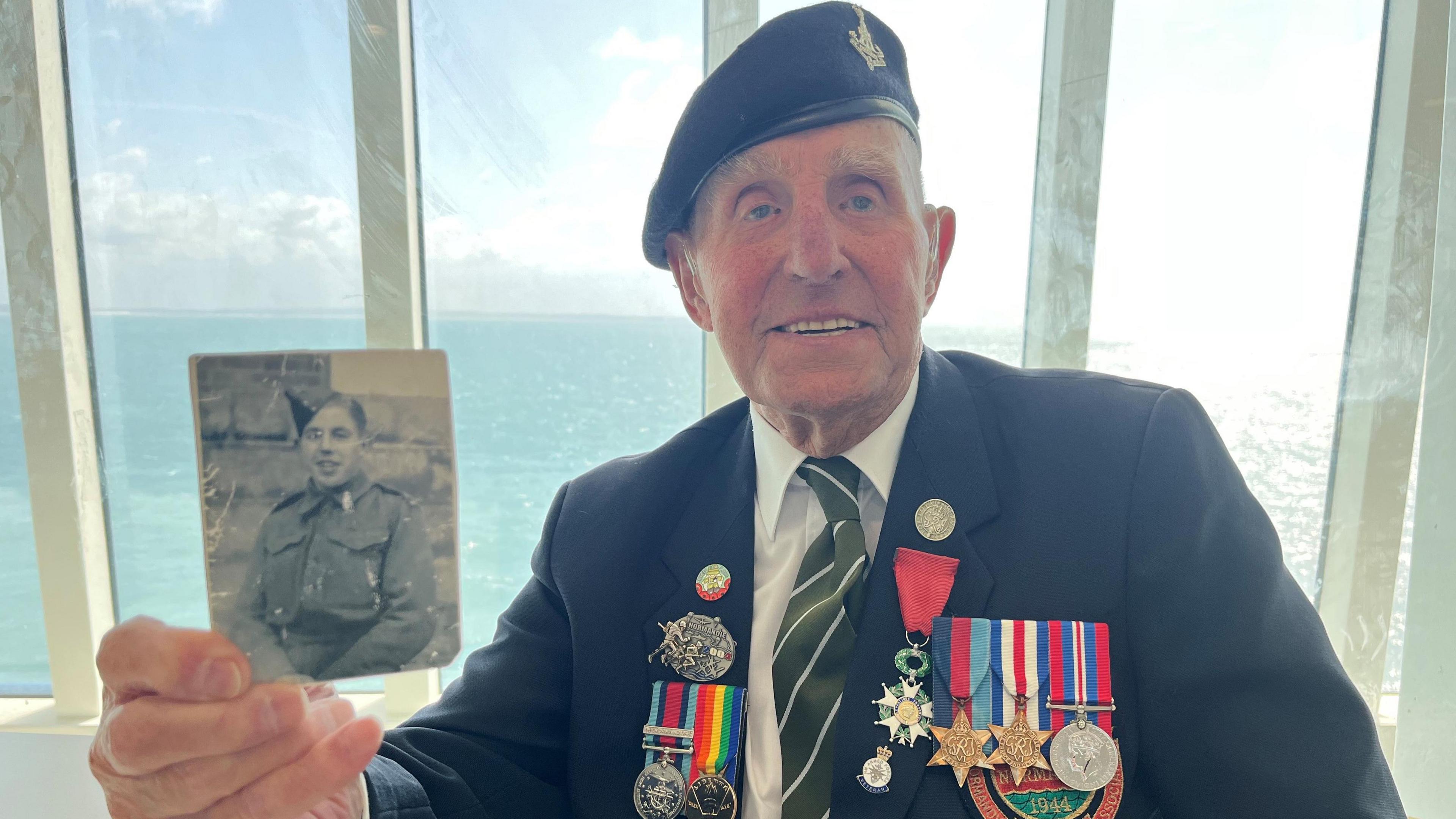 A old man wearing military medals smiles as he holds a black and white photo of a young man in uniform. The sea is in the background.
