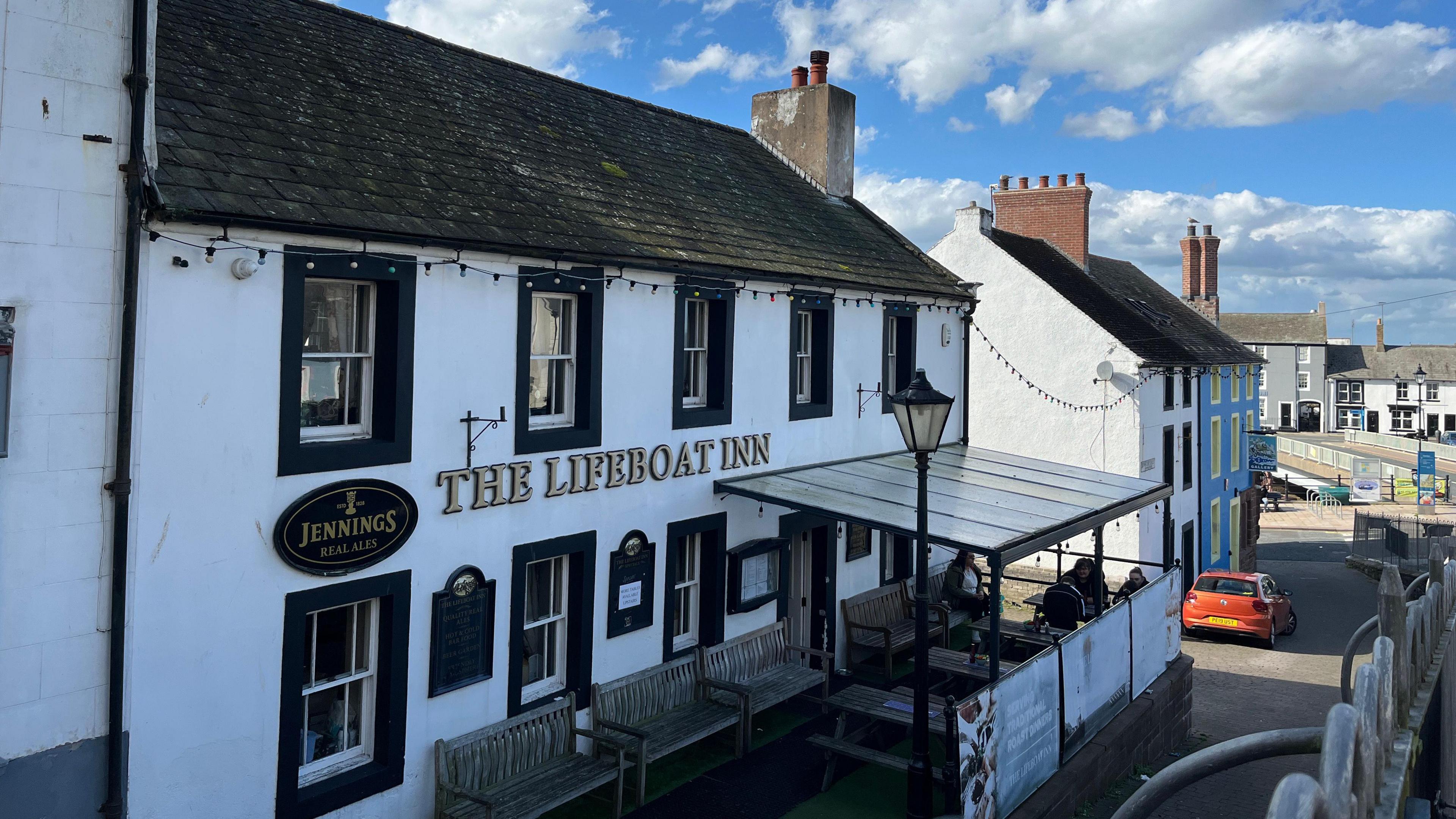 The Lifeboat Inn at Maryport. The two-storey white building has black details around the windows and door. The name is on the front of the building in golden lettering. A raised area at the front of the pub is covered by a metal framed canopy with clear sheeting.