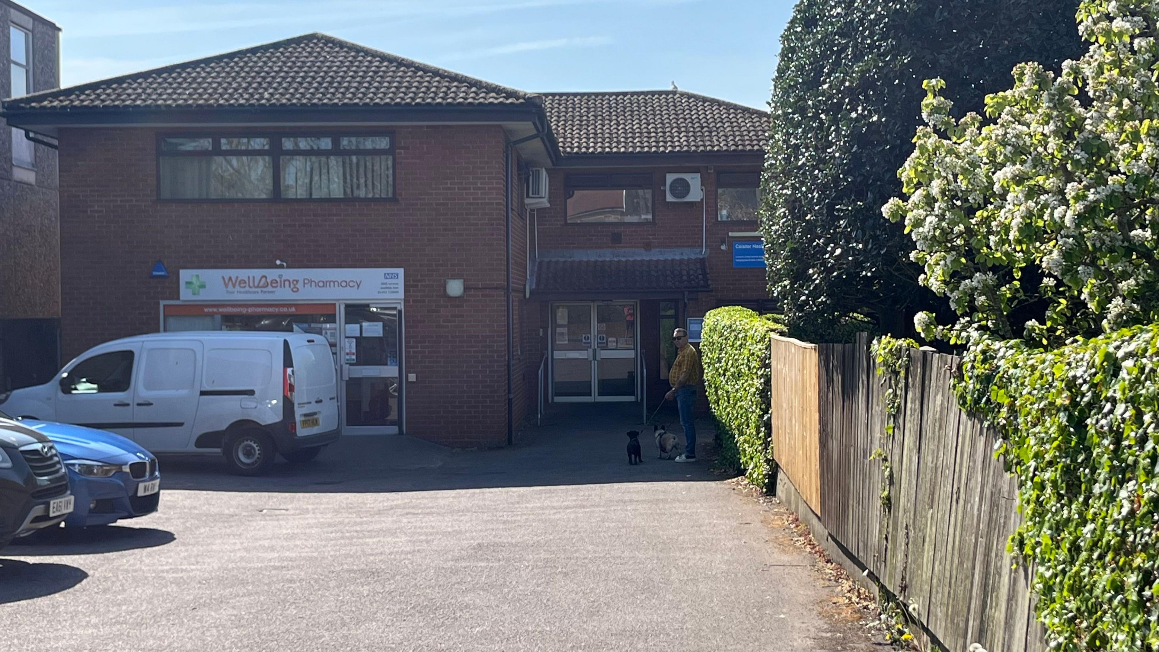 A modern two-storey red brick building with a man standing in front with two dogs. There is a pharmacy at the front of the building, which stands behind a car park with a van and cars.
