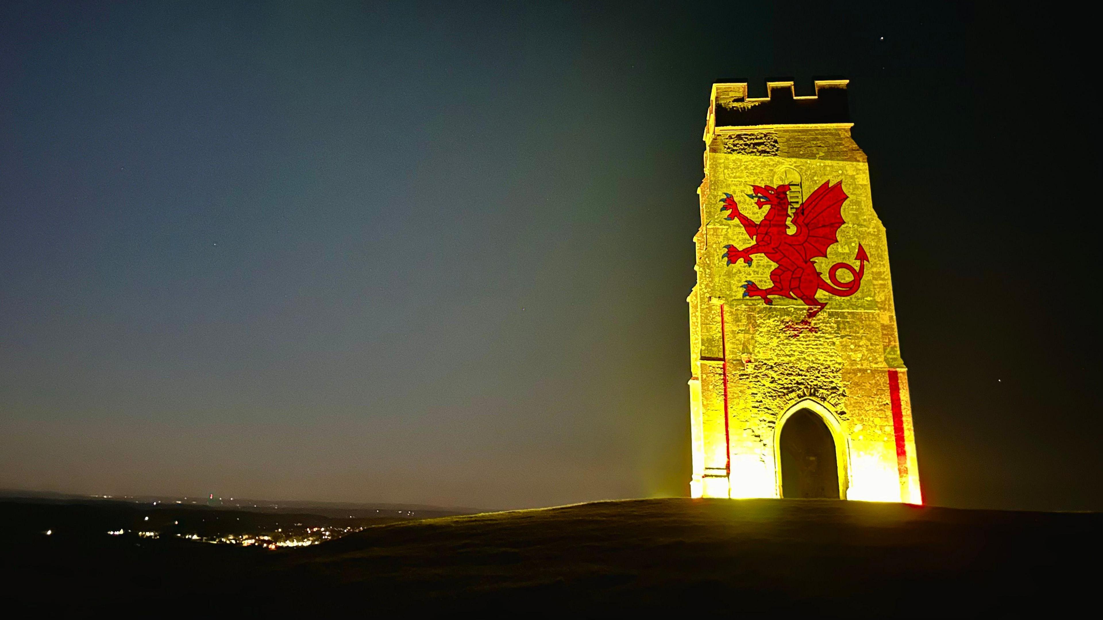 Glastonbury Tor is lit up yellow with the red dragon of the Somerset flag displayed in the middle. It is night time.