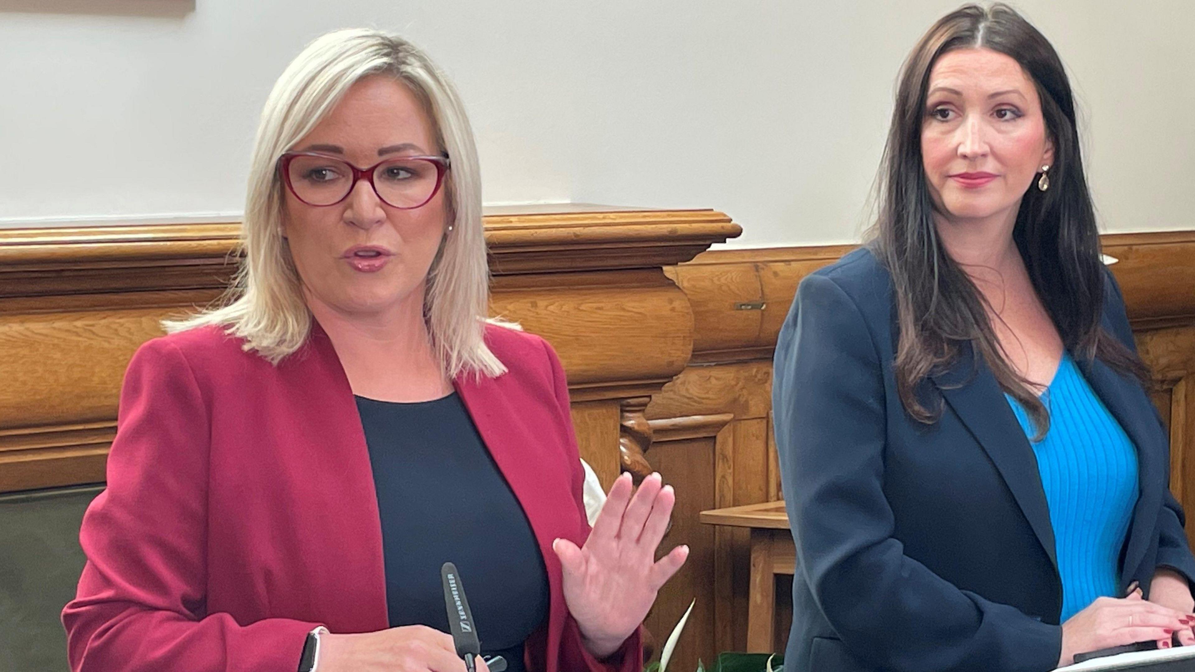 Michelle O'Neill and Emma Little-Pengelly standing behind podiums with microphones. Michelle O'Neill is speaking. They are standing in a room with wood panelling in the background