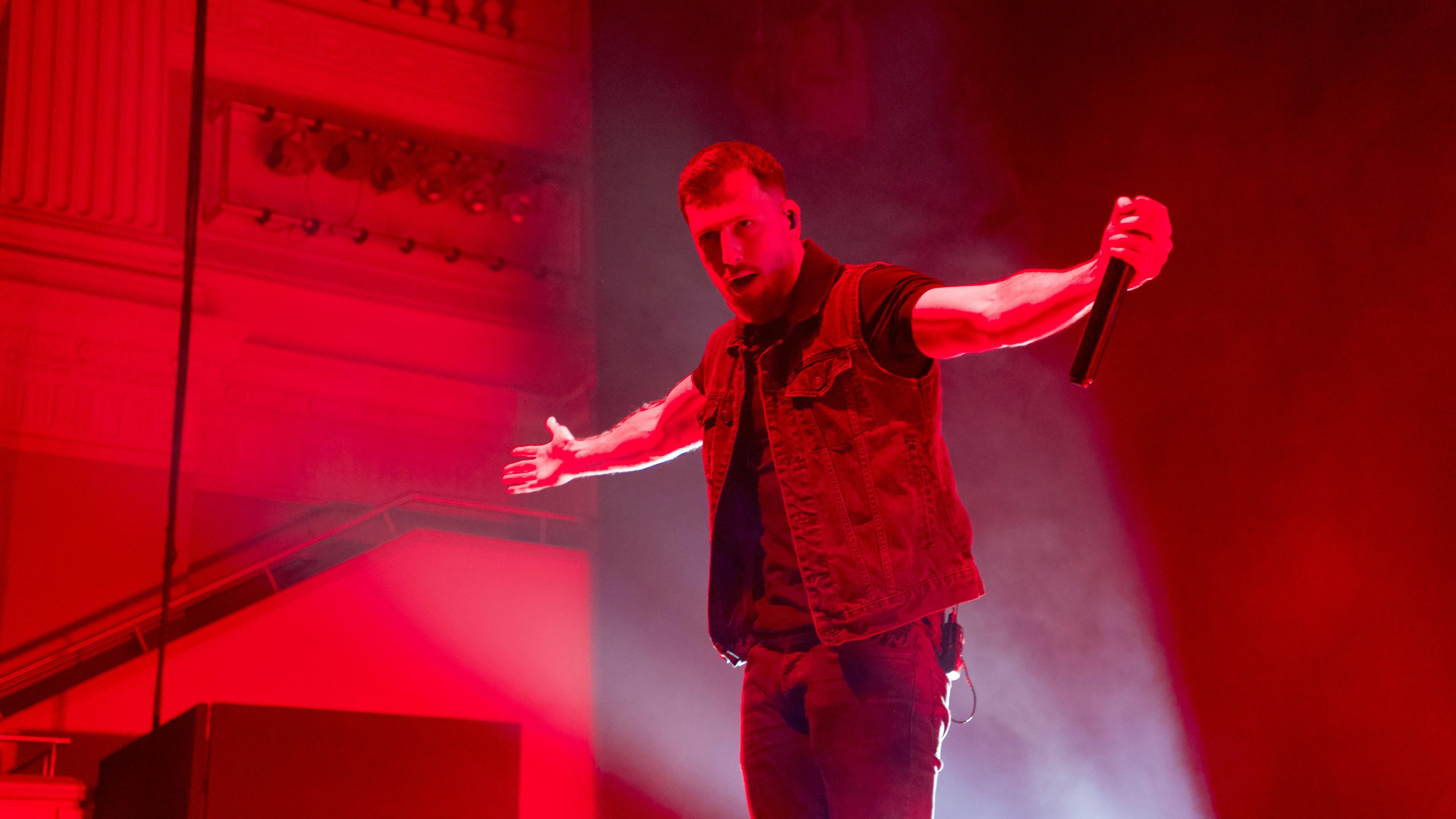 A lead singer on stage with arms outstretched taking the applause of fans. The stage lighting is read and he's wearing a denin waistcoat