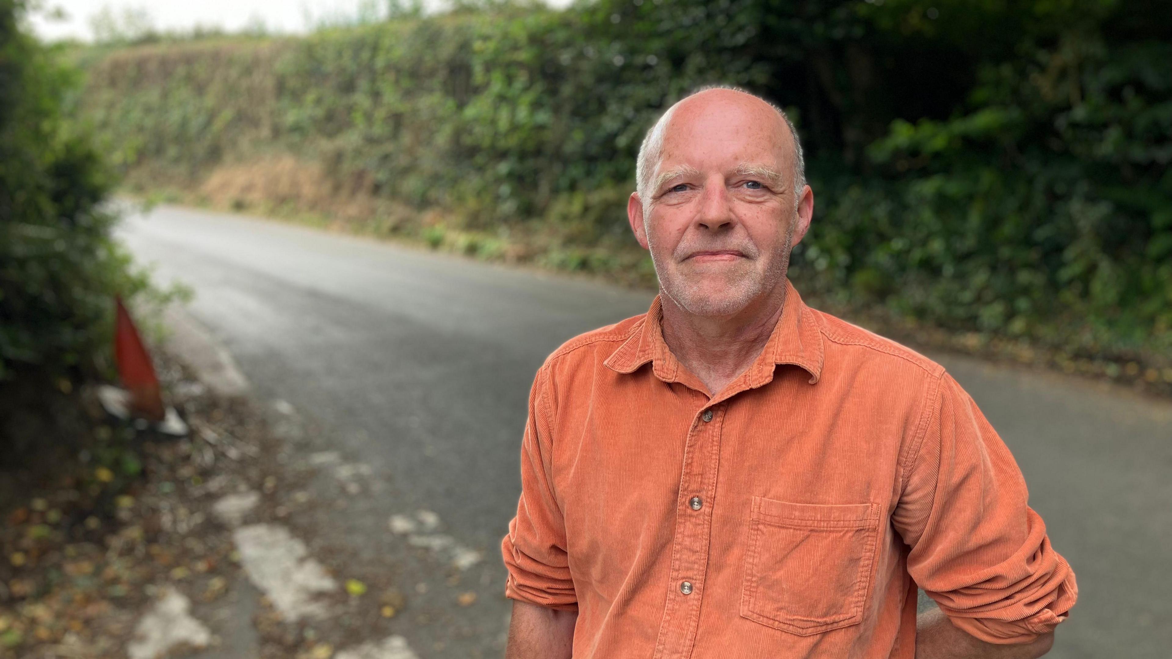 A man in an orange shirt stands in a country lane. There are thick, high hedges behind him. 