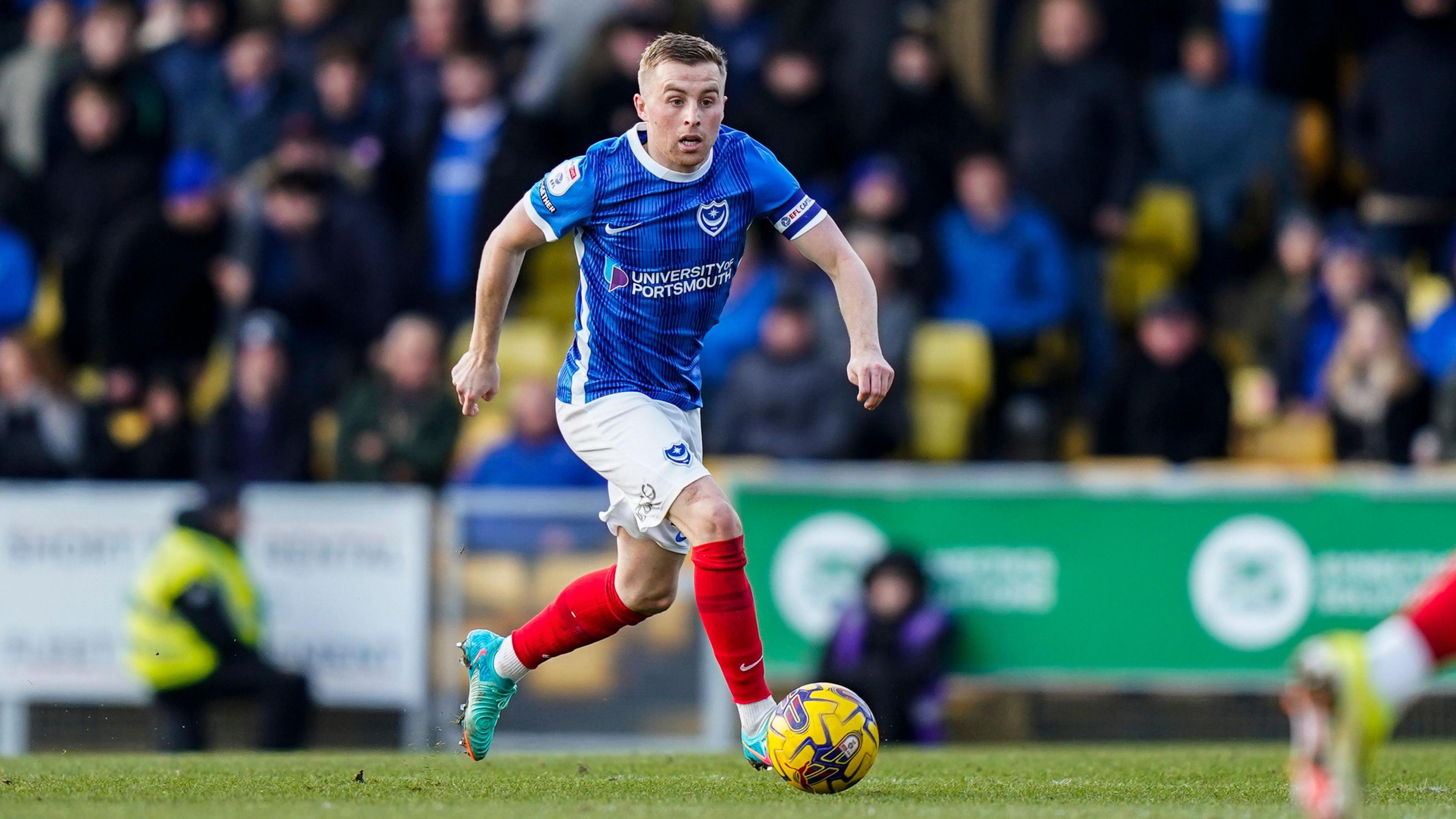 Joe Morrell wearing Portsmouth blue kit running on a pitch with the ball at his right foot during a match in January 2024