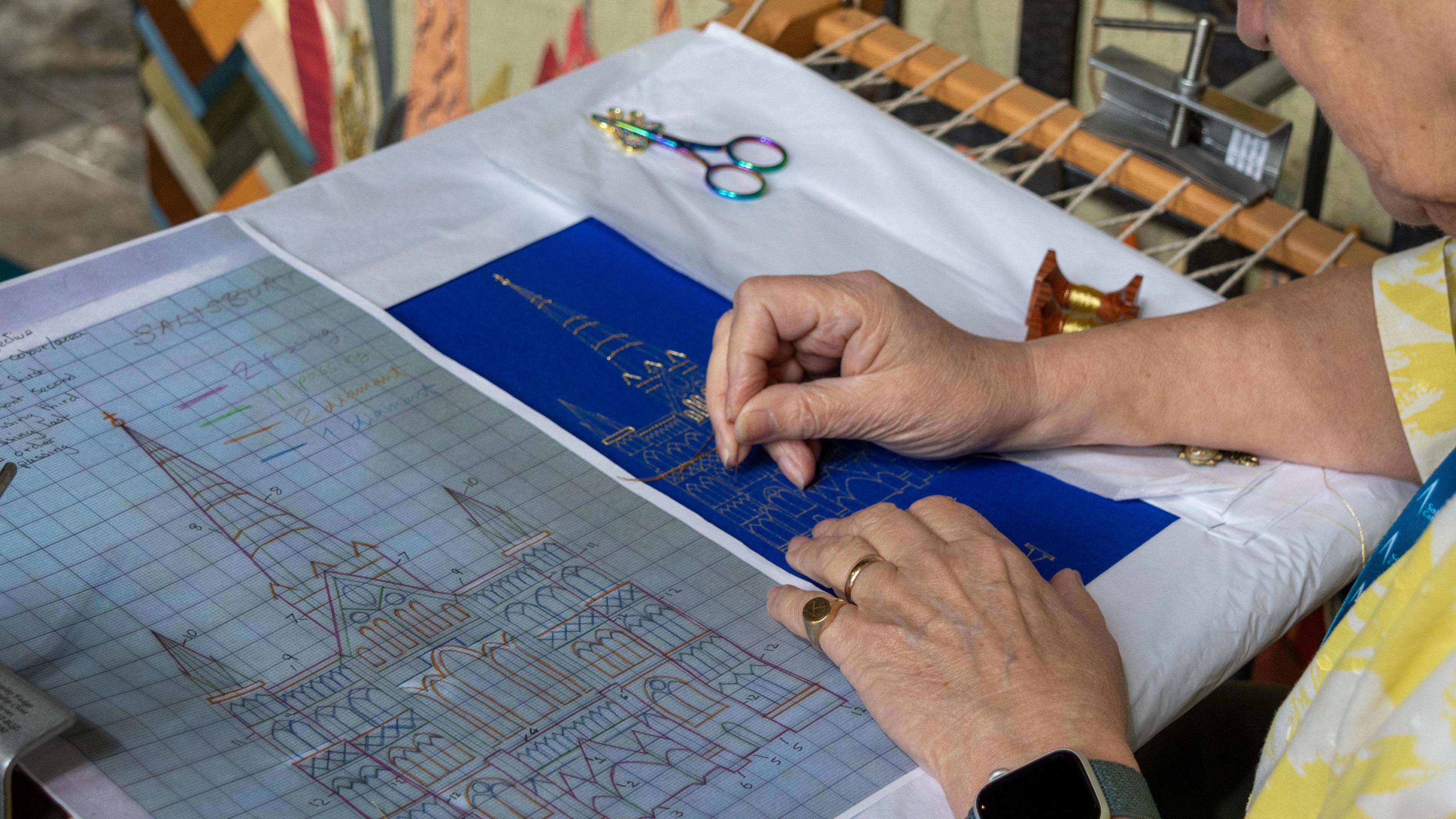 A woman's hands stitching onto a white piece of cloth. An image of a cathedral is on the left hand side as a guide.