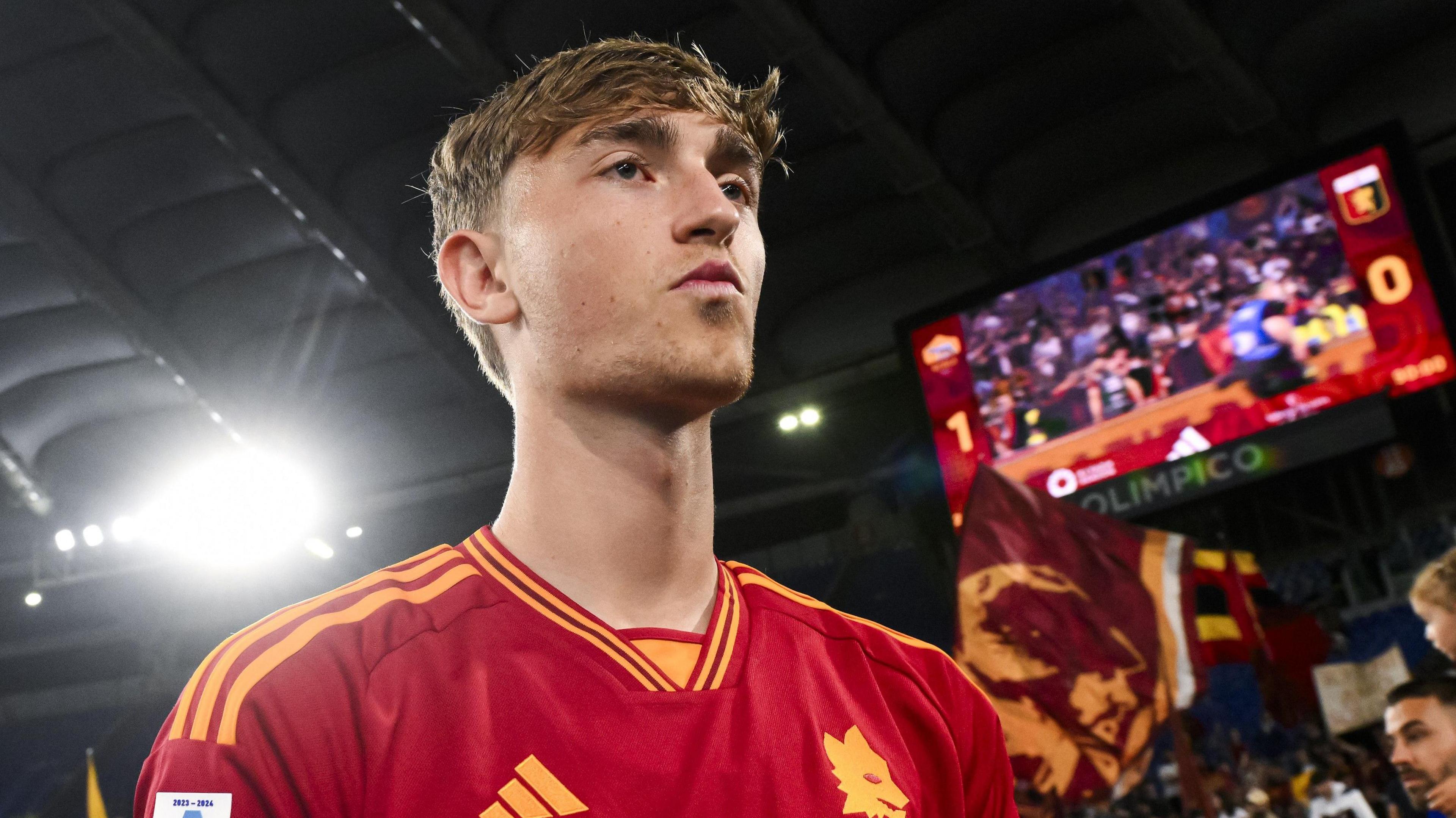 A picture from slightly below Dean Huijsen, wearing Roma's red shirt. In the background one floodlight glares behind his right shoulder and behind his left is a big screen showing the crowd