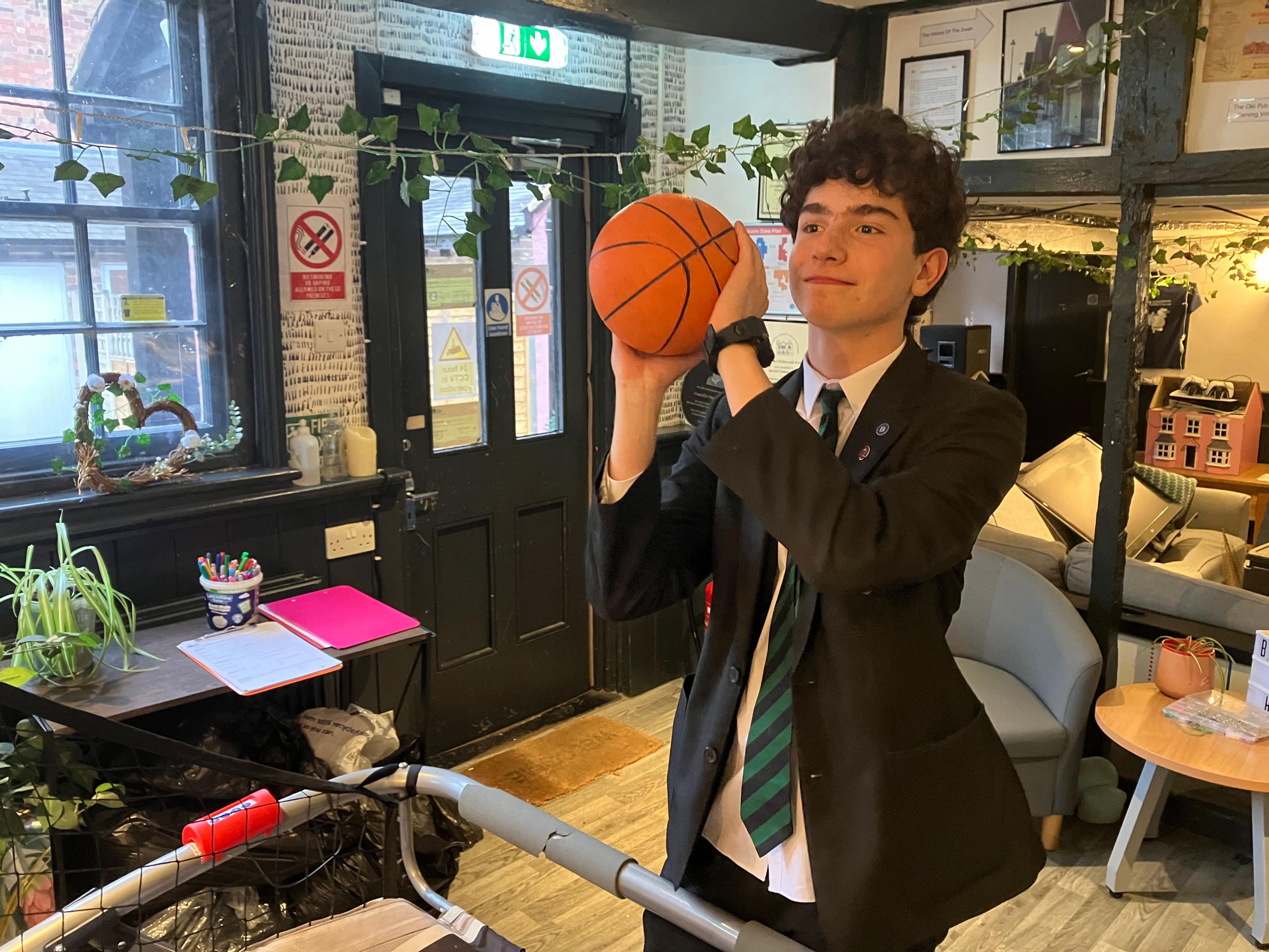 An image of a teenage boy with black curly hair. He has a basketball in his hand as if he is about to take a shot, and he is wearing a black blazer and green and black striped tie.