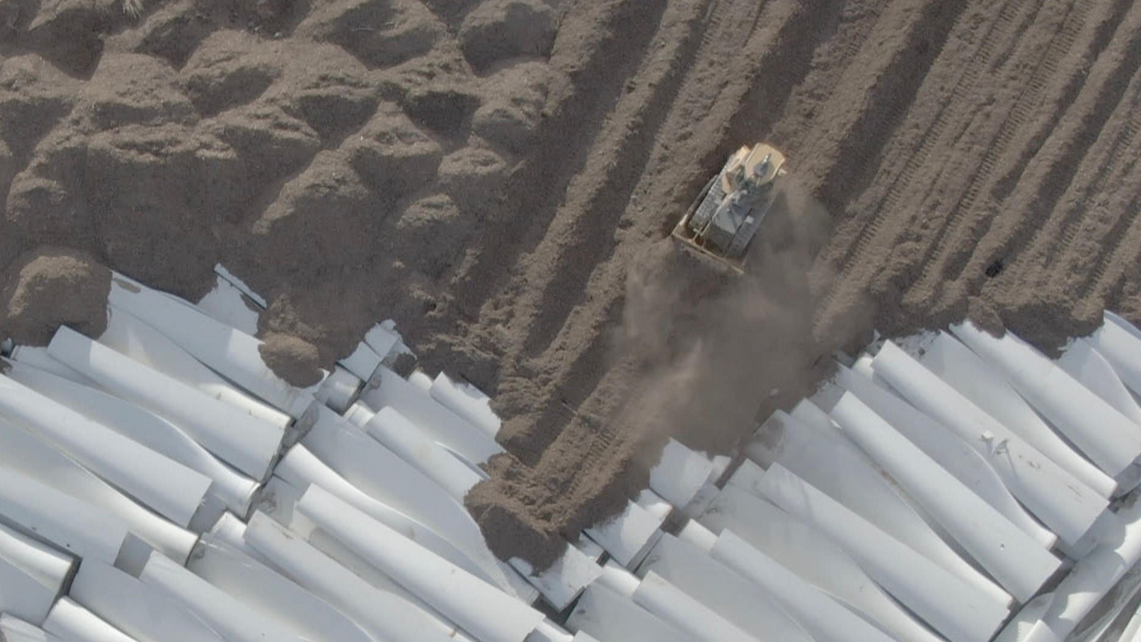 A bulldozer is seen from drone footage piling soil on top of dozens of old wind turbine blades. 