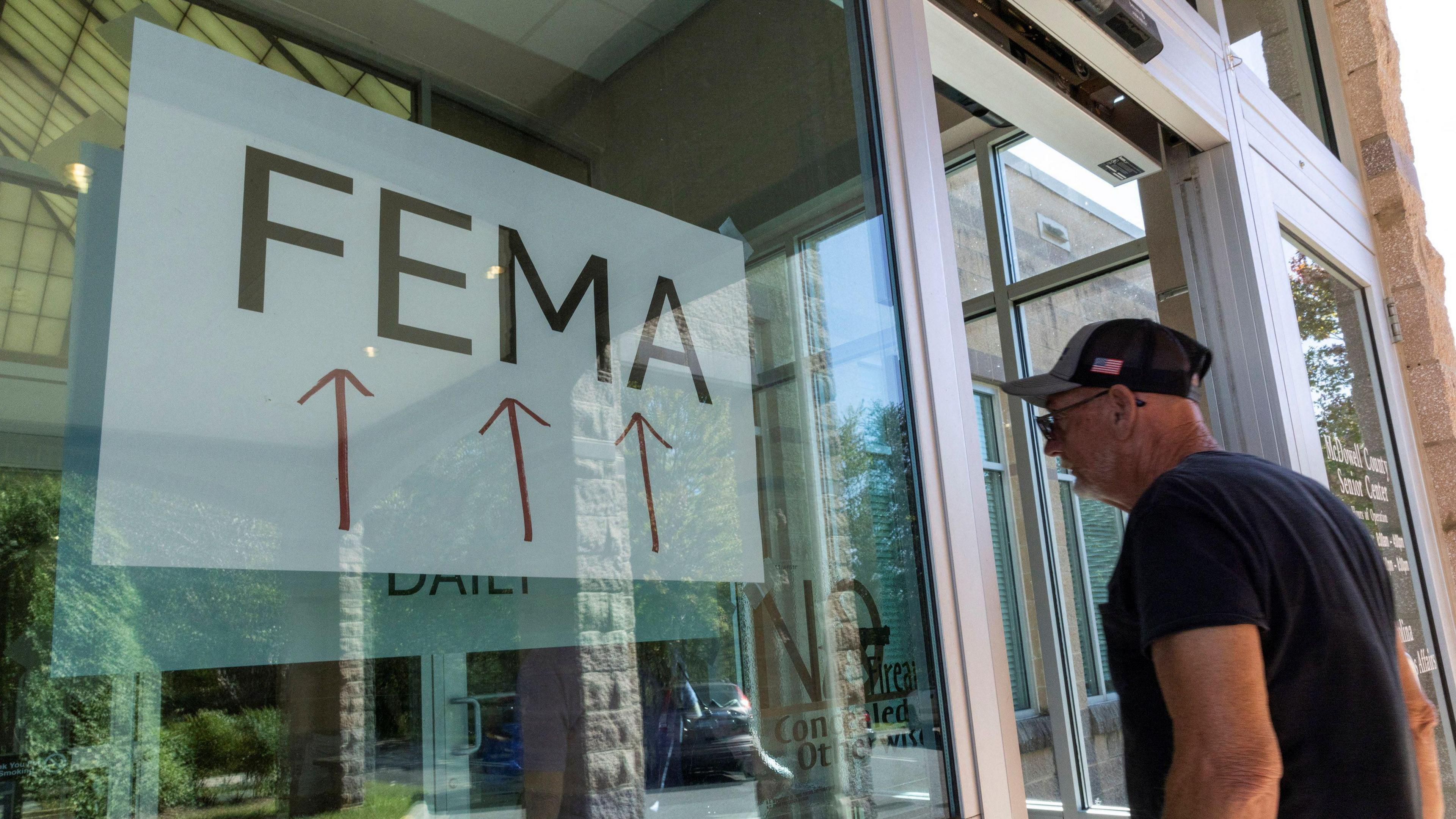 An elderly man wearing a baseball cap enters an office with a large sign saying FEMA and three red arrows 