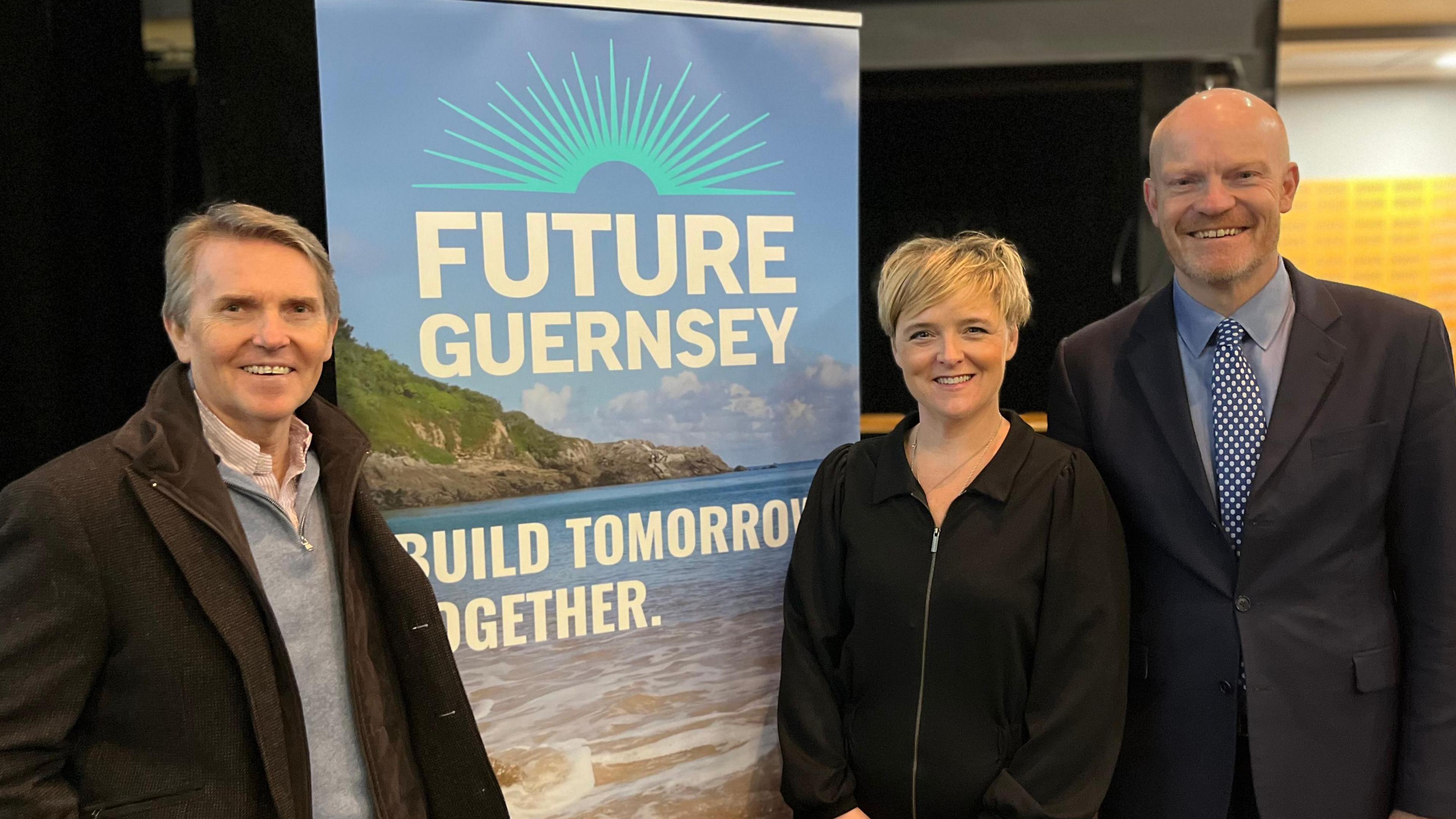 From left to right, a man with grey and black hair wearing a brown coat and a grey jumper over a pink shirt smiling at the camera, standing next to a banner that says Future Guernsey, Build Tomorrow Together. Next to the banner on the right is a woman with short blonde hair smiling, wearing a black top, on her right is a bald man smiling wearing a black suit and a blue  and white tie with a blue shirt. 