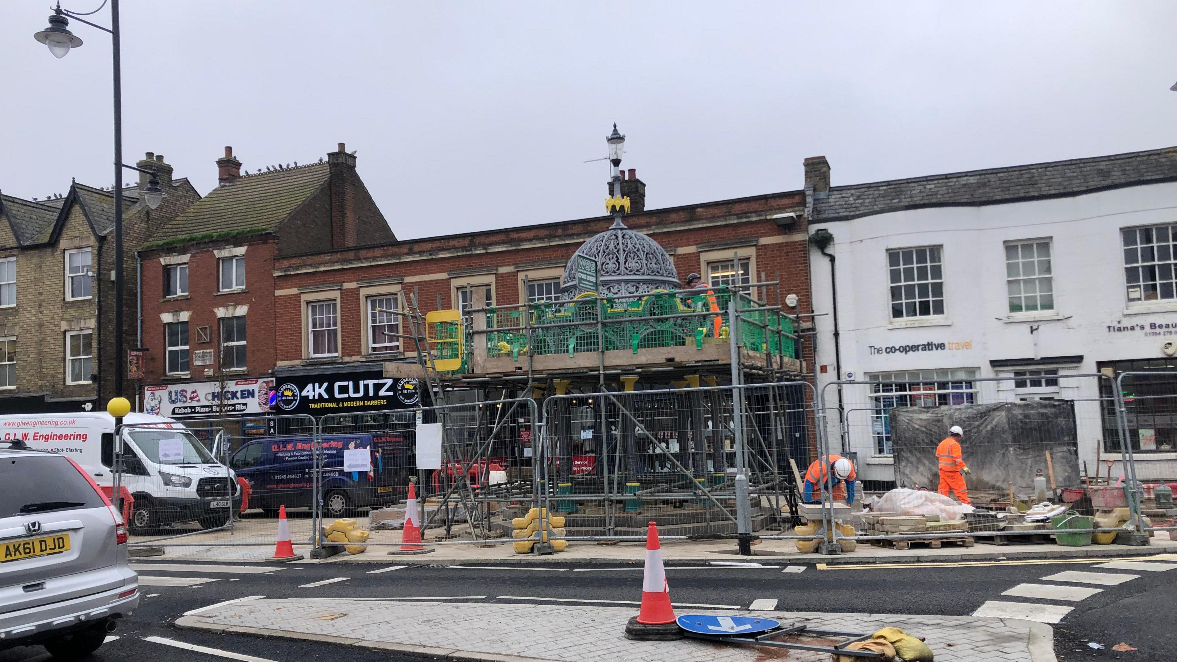 The Fountain structure with the grey dome, with green and yellow trim visible but the bottom section covered in scaffolding with workmen around it. Behind it are various shops on Broad Street in March.