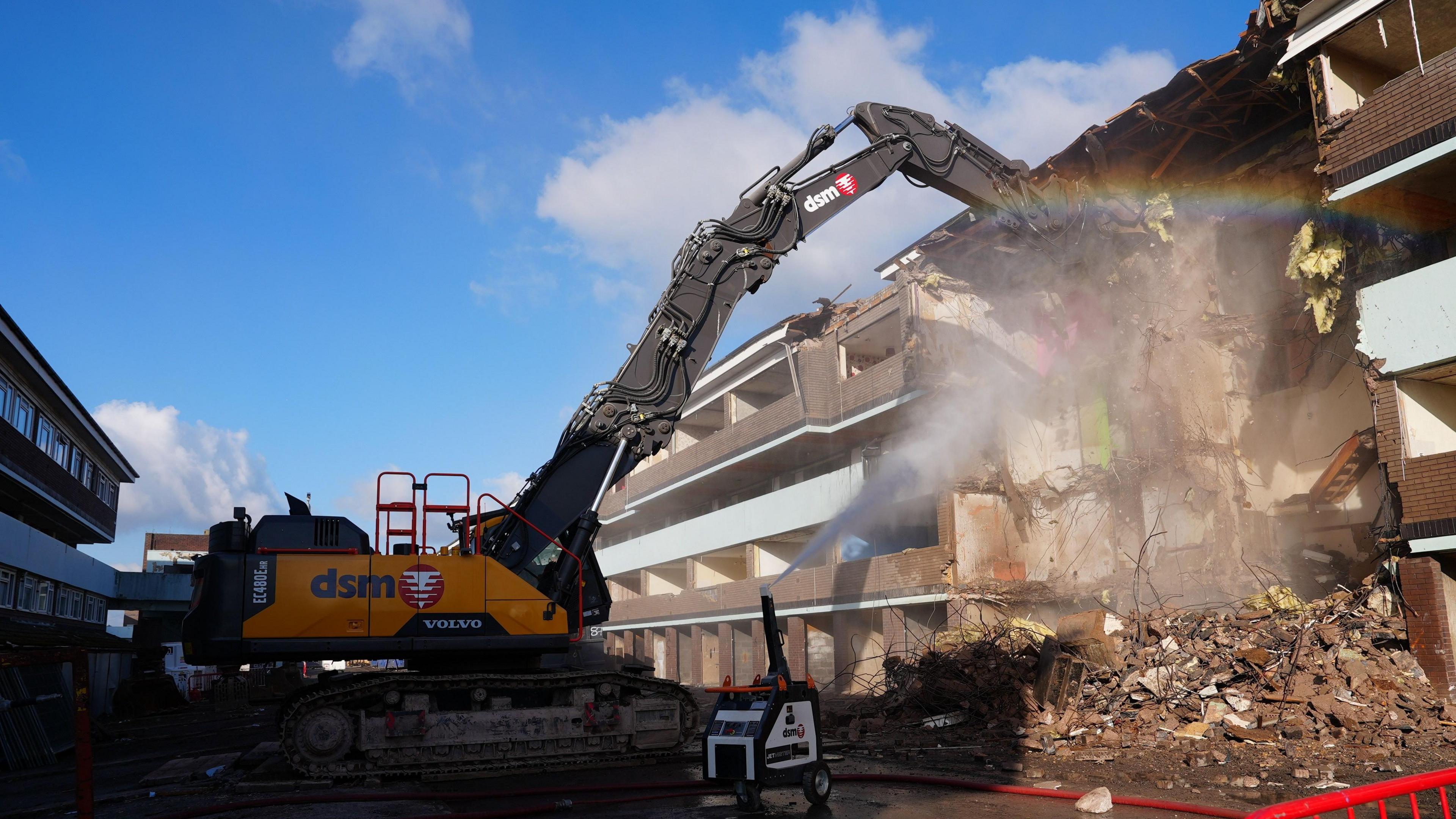 A digger begins demolishing a block of maisonettes. The sky is bright blue and a  rainbow effect is seen near the digger.