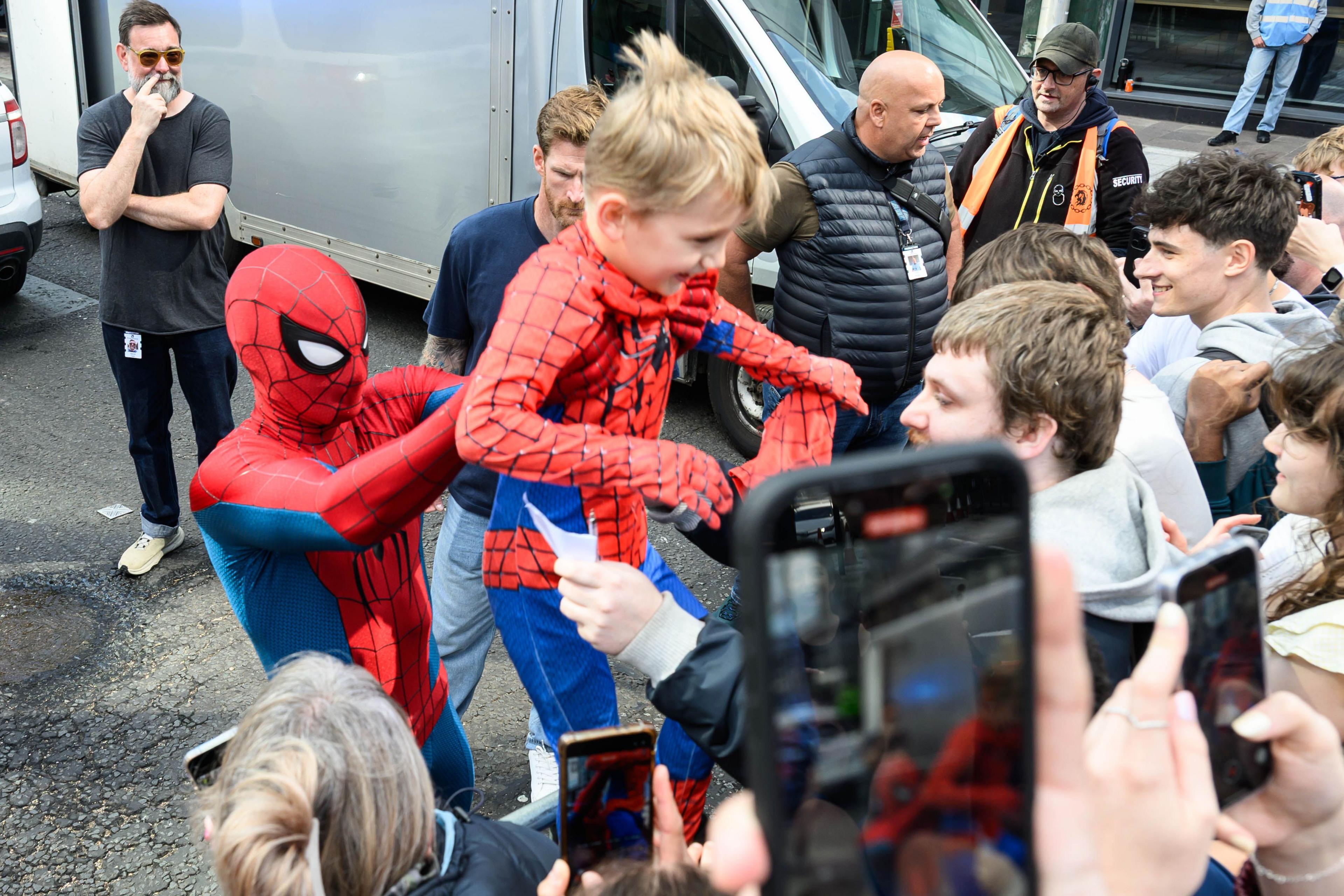 Spider-Man lifting a young lad over a barrier for a photo. The boy is wearing a Spider-Man costume and is smiling. The crowd is enjoying the interaction.