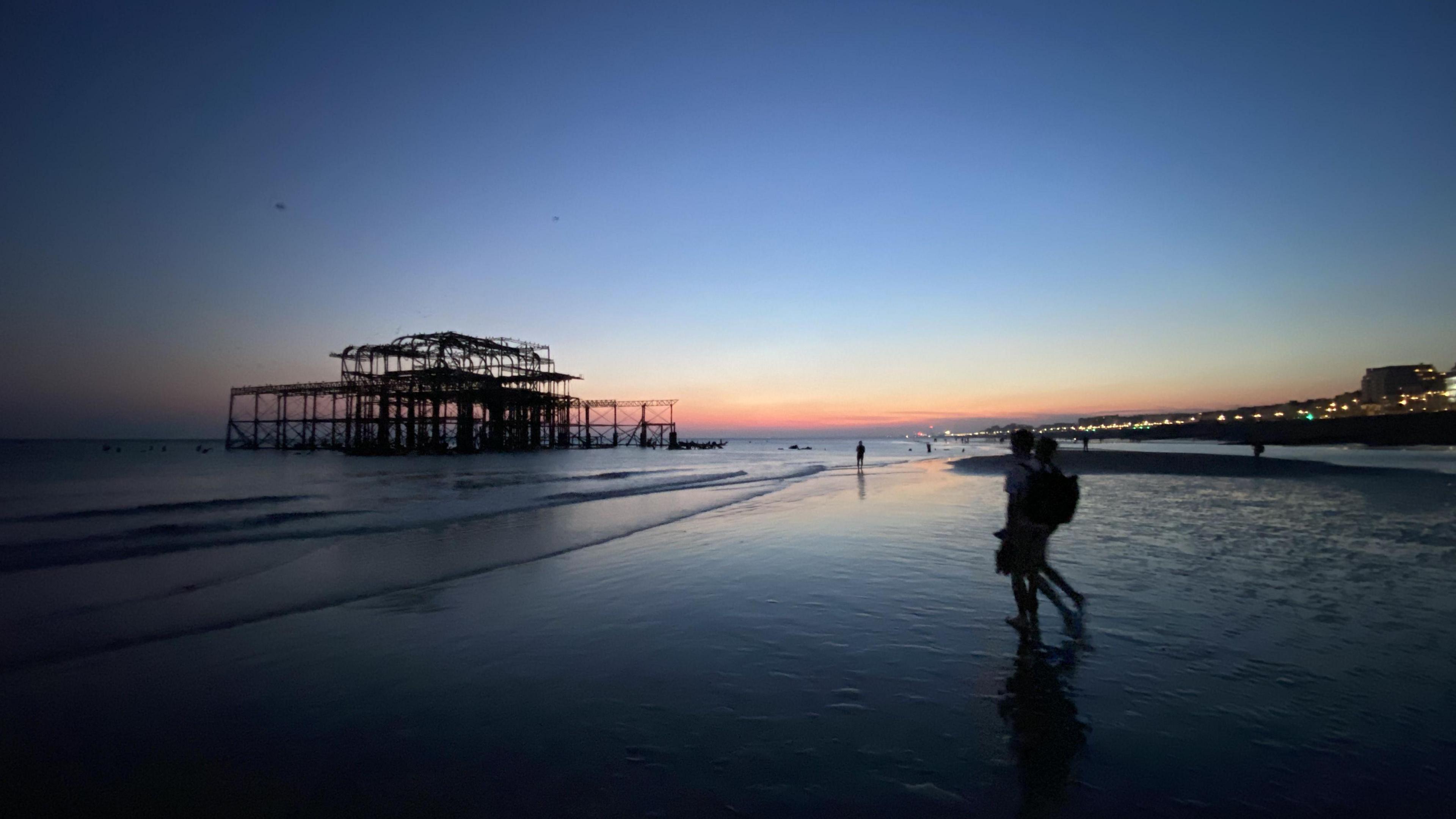 Sunset at low tide on Brighton seafront. To the left is the sea and the West Pier, a silhouette in front of a blue and orange sky. There are two people walking on the right