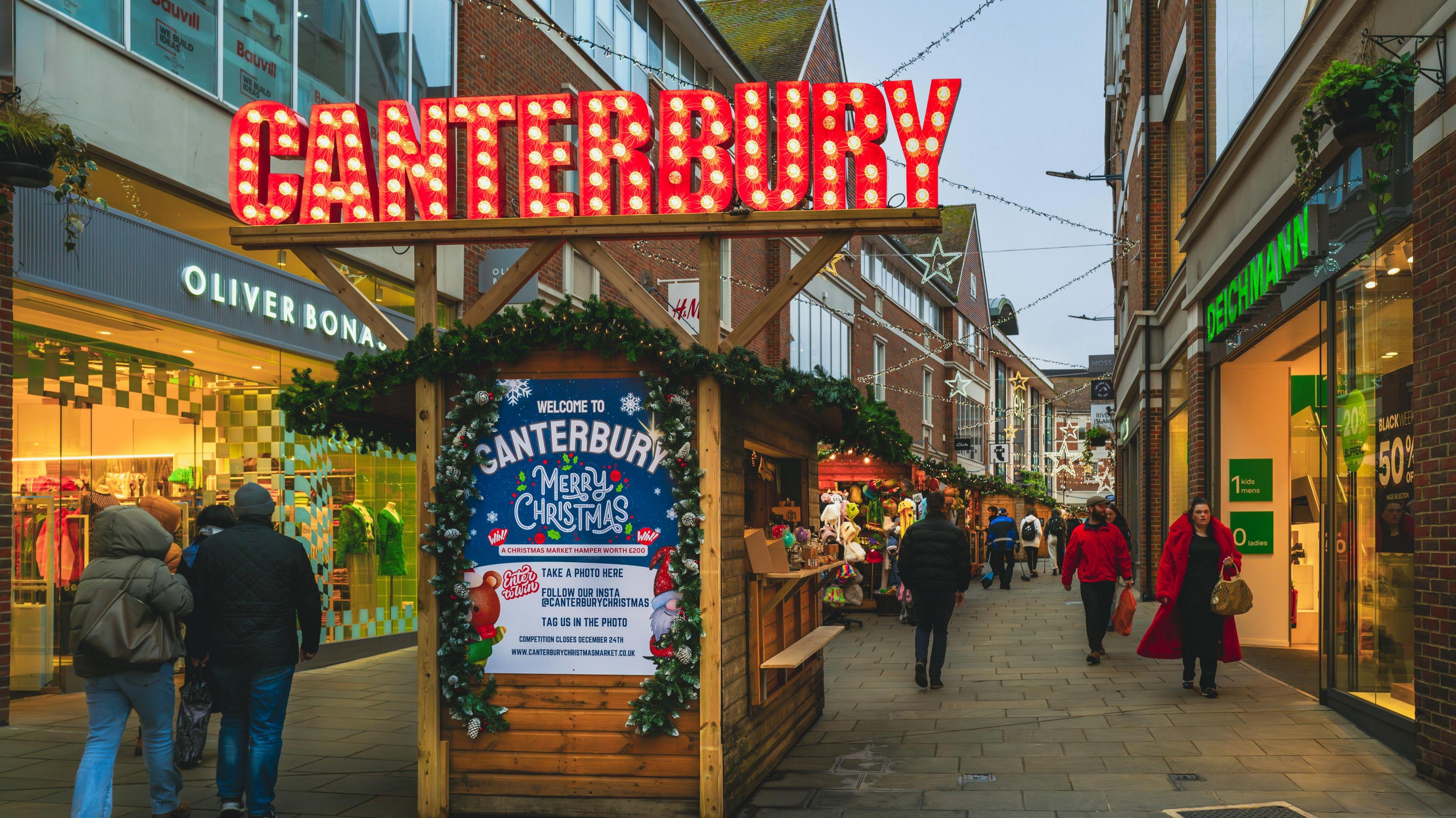 A stall decorated with a large illuminated sign saying "Canterbury" on top of a wooden hut in a city street decked out for Christmas, as shoppers walk past.