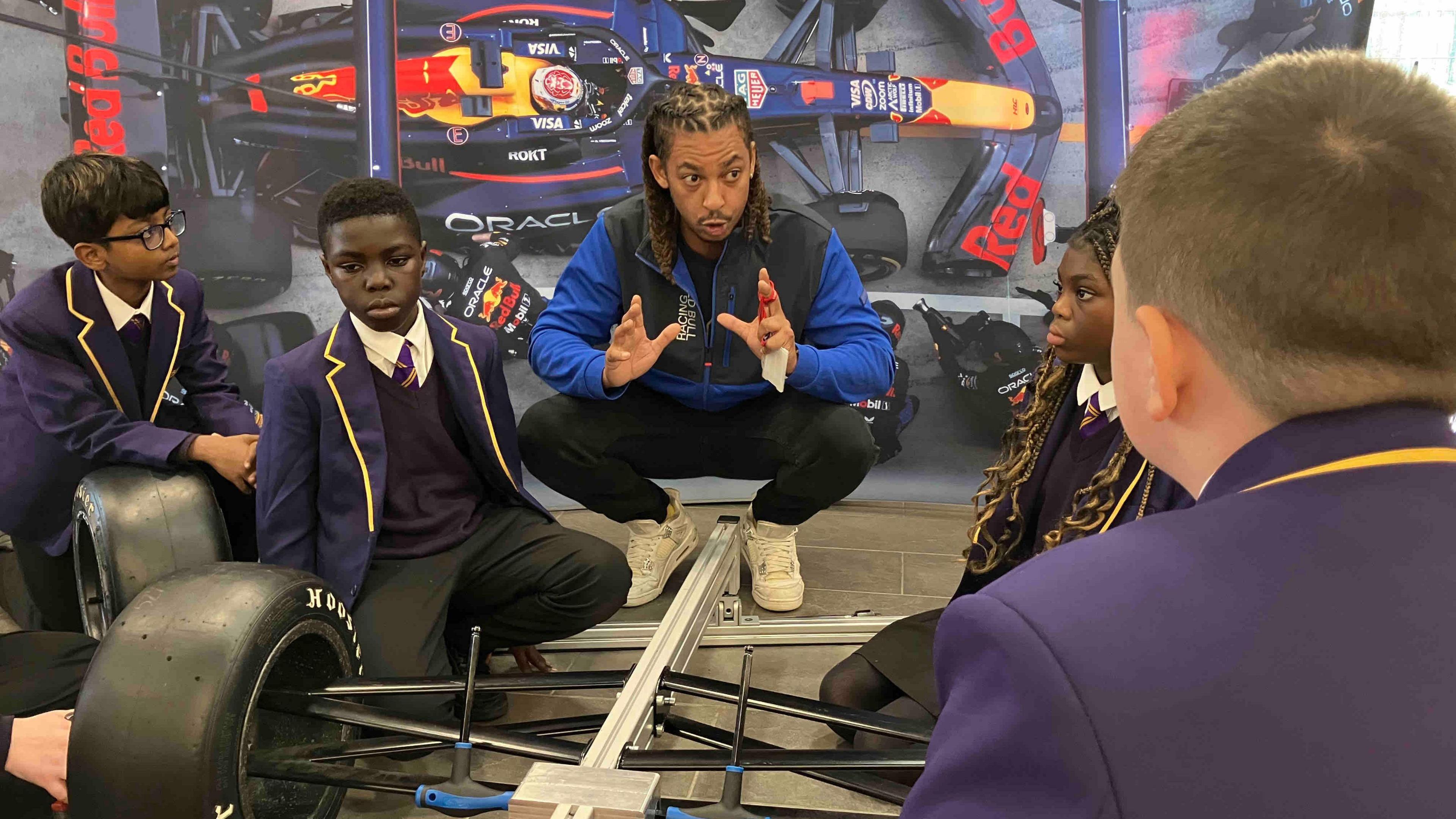 A man is crouched down and speaking to children dressed in purple school uniforms. There is a picture of an F1 car behind him and a mock-up car in the foreground.