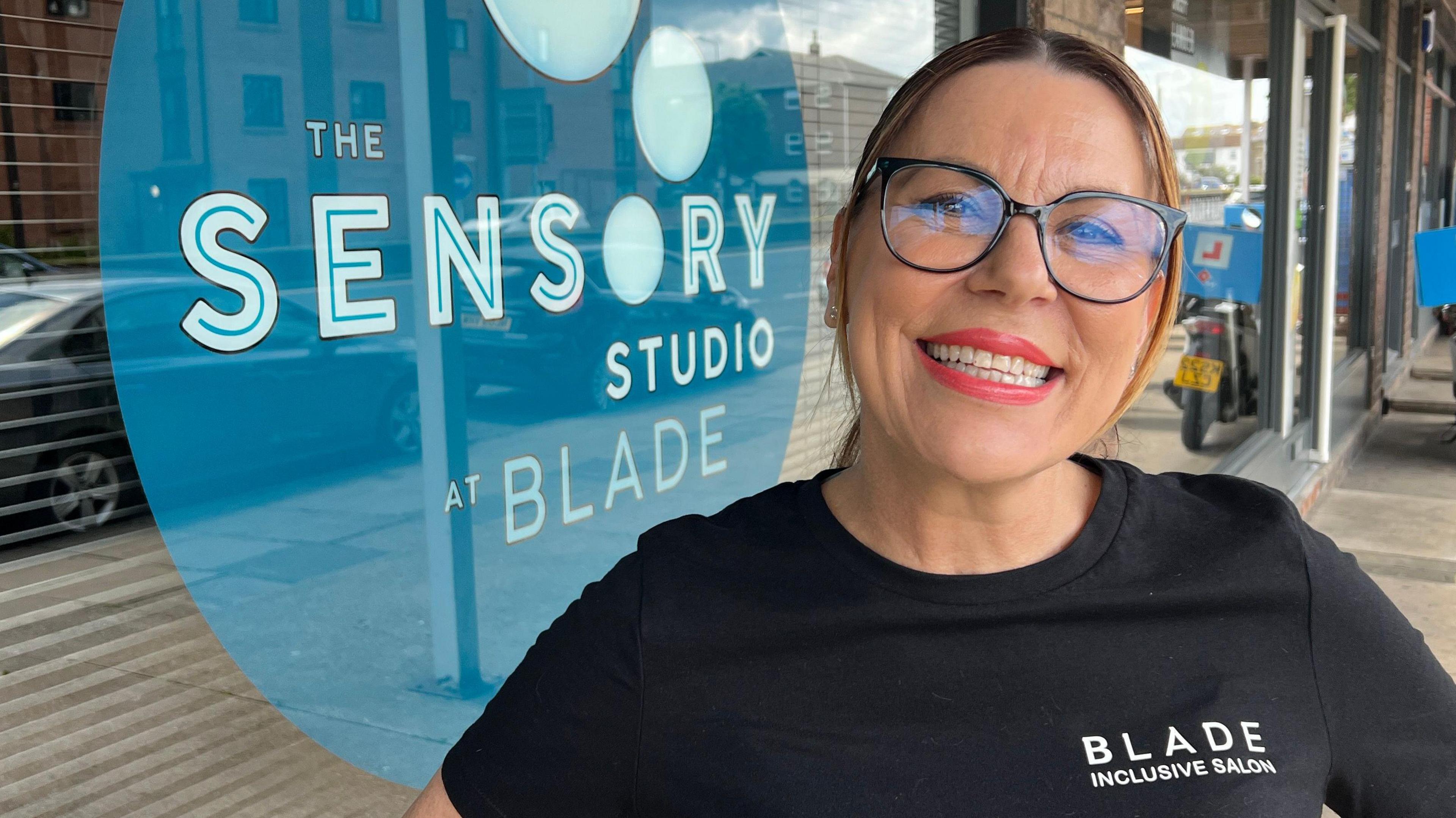 A head and shoulders image of a woman wearing a black top with the words Blade Inclusive Salon on it. She is looking into the camera.