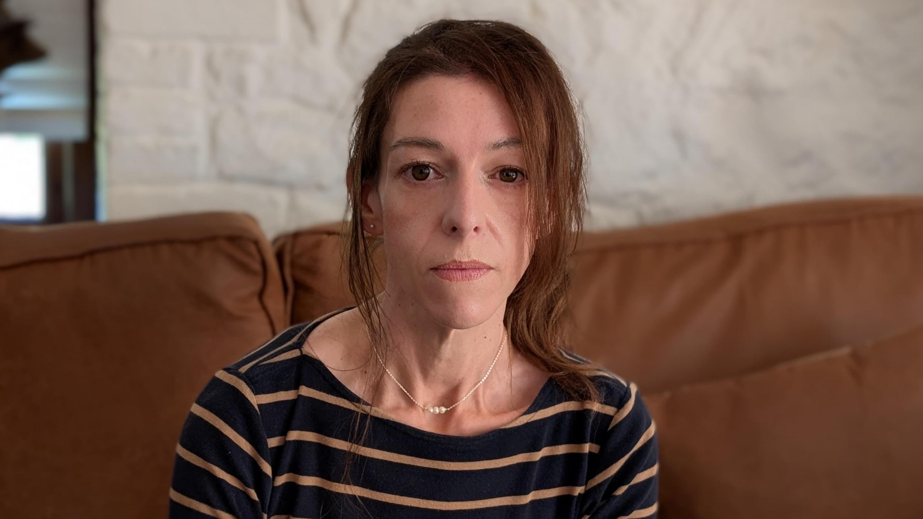 Emma Simmonds is sitting down on a brown sofa. She is looking directly at the camera and is wearing a navy and brown striped long-sleeve T-shirt. She has brown hair, pulled back in a ponytail.