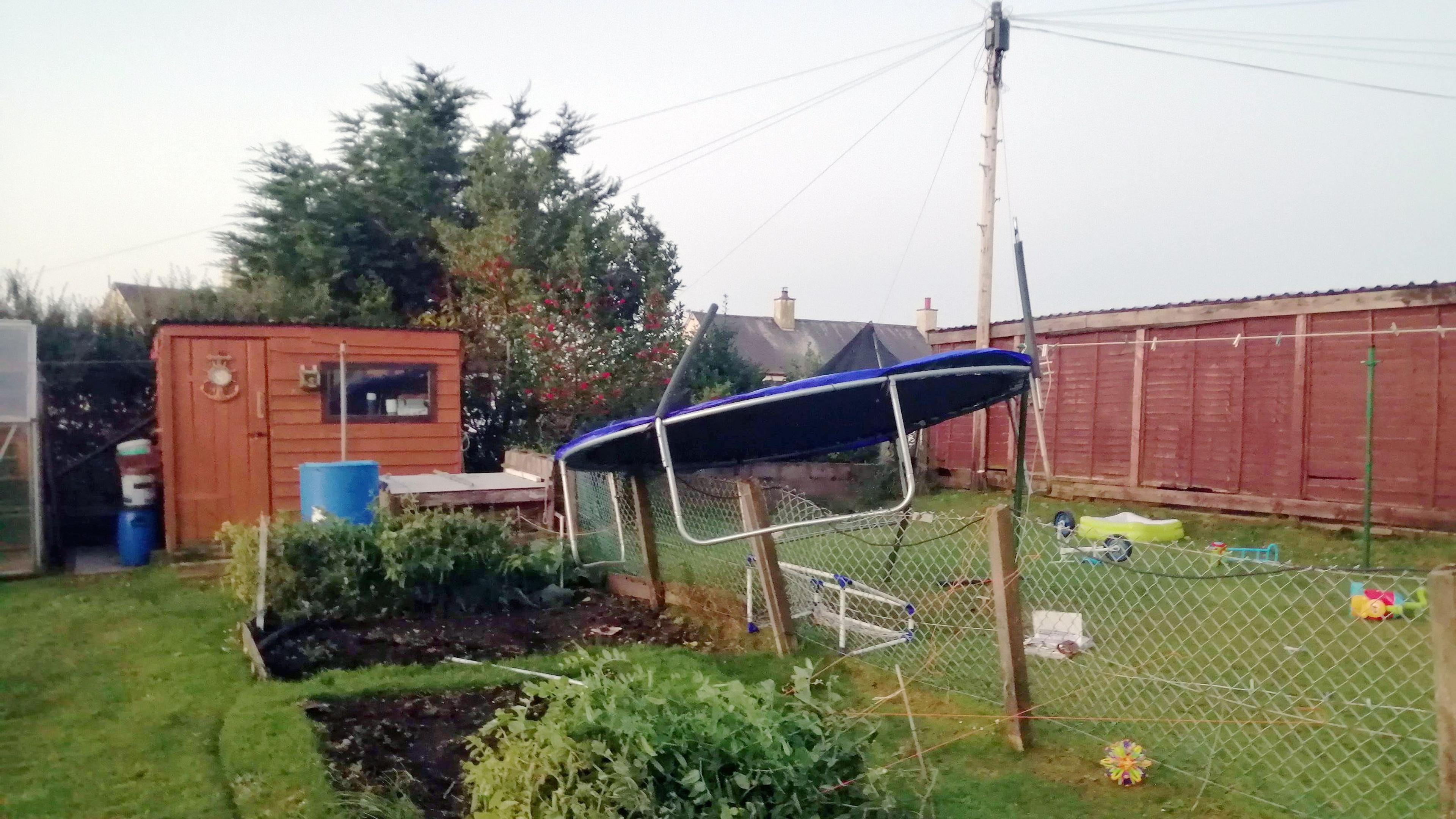 A trampoline in a garden sitting on top of a chain link fence with other garden toys strewn over a lawn with a hut in the background