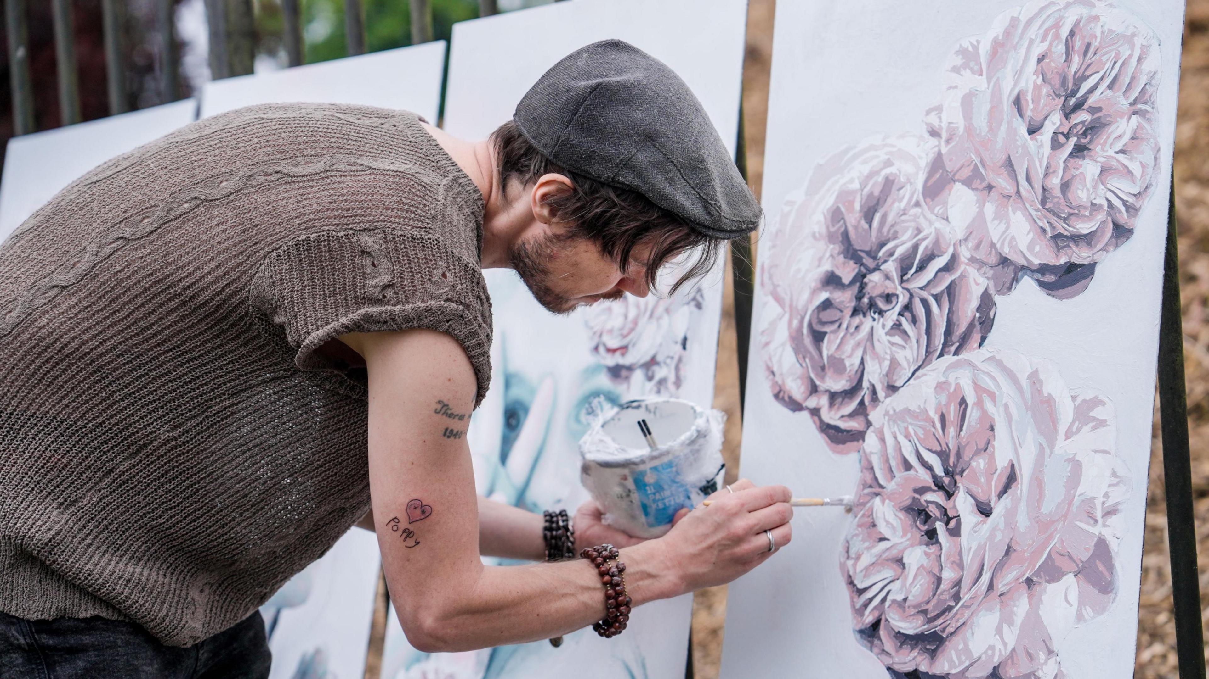 A man with dark hair wearing a grey cap paints light pink flowers onto a canvas. 
