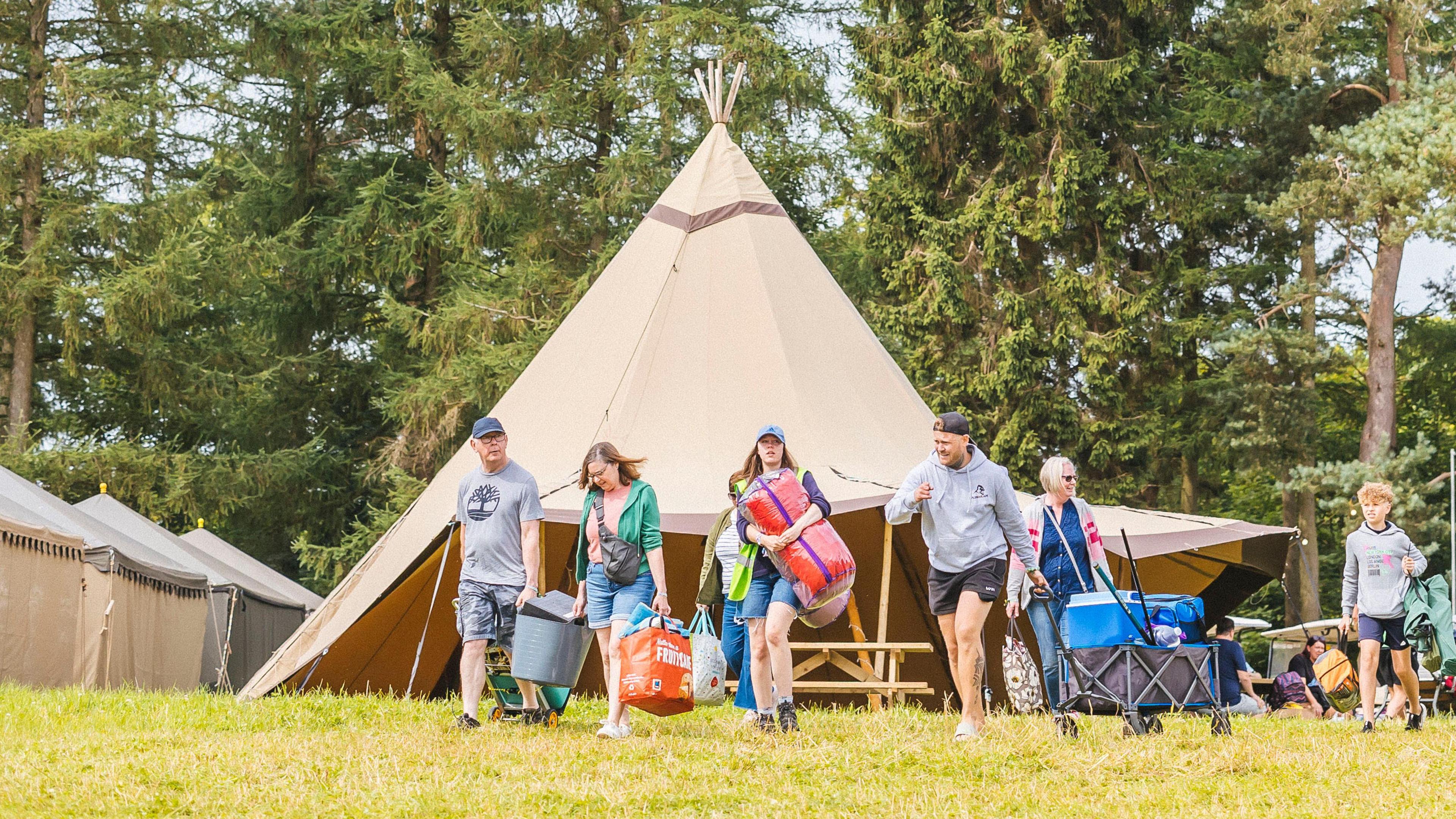 A family walk across a field carrying bags in front of a tipi tent.