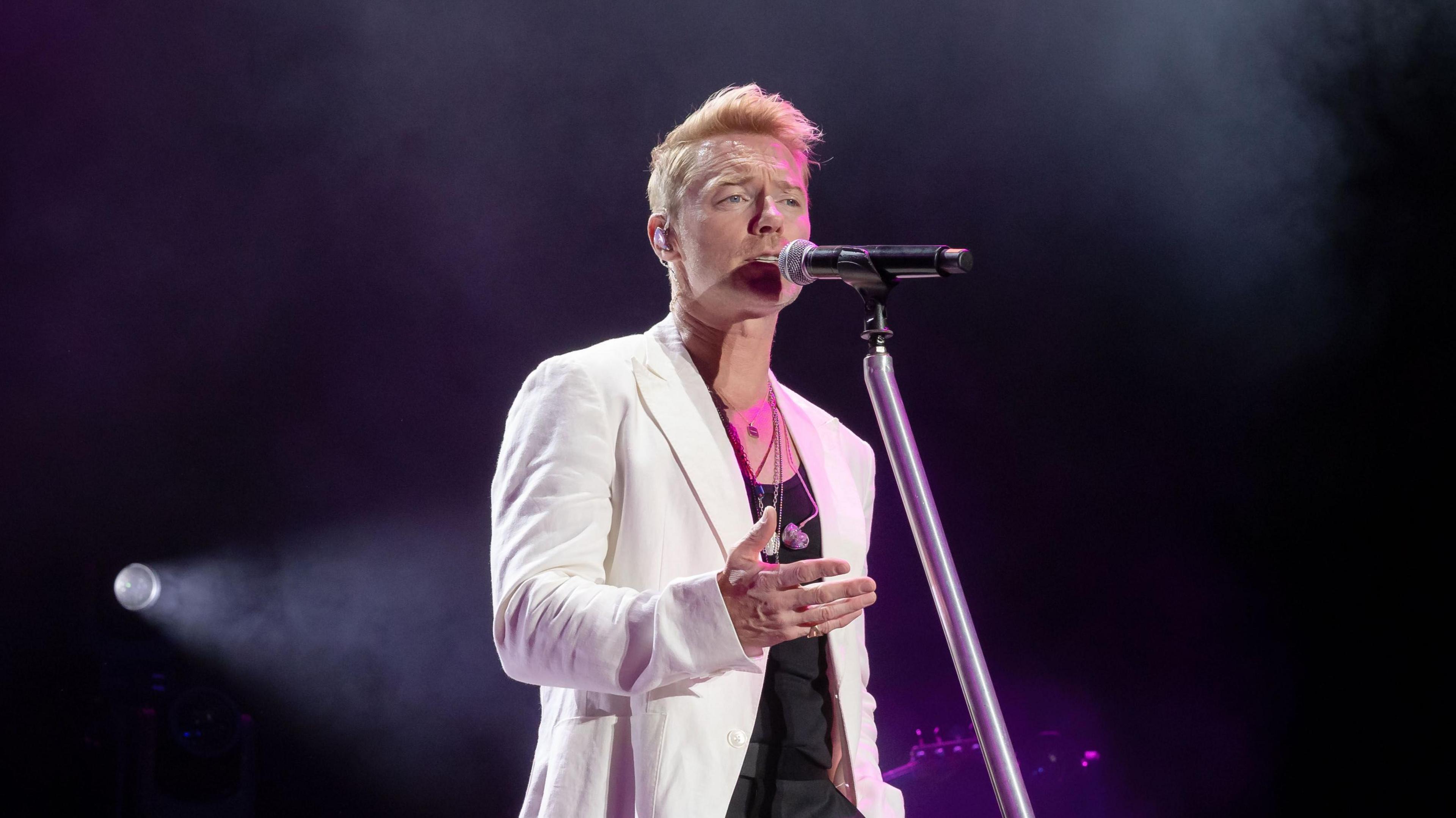 Ronan Keating stands by a microphone stand in front of a dark background with a spotlight. He wears a white blazer and black suit trousers.
